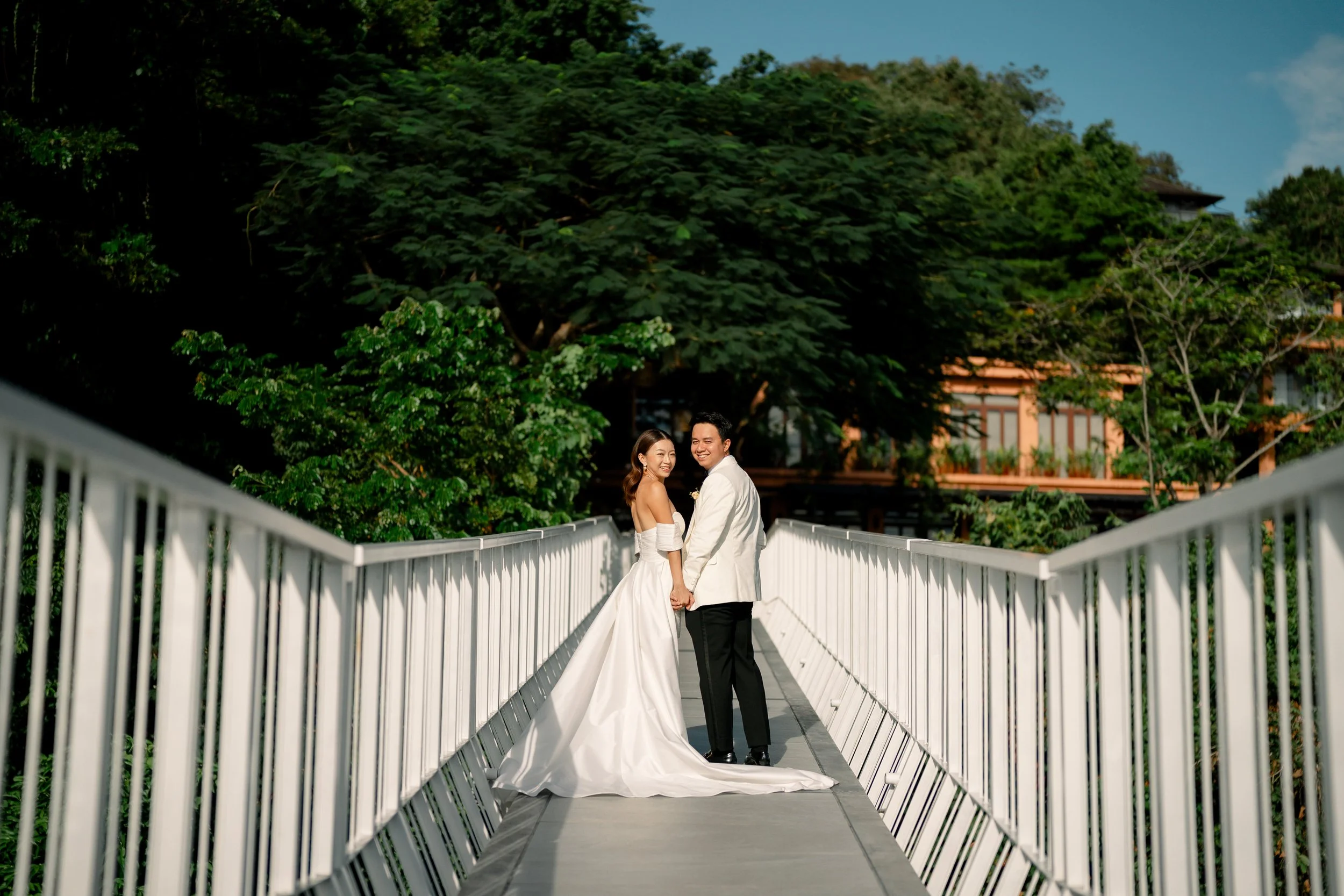 A bride and groom in wedding attire standing on a white bridge surrounded by lush greenery and trees, with a building partially visible in the background.