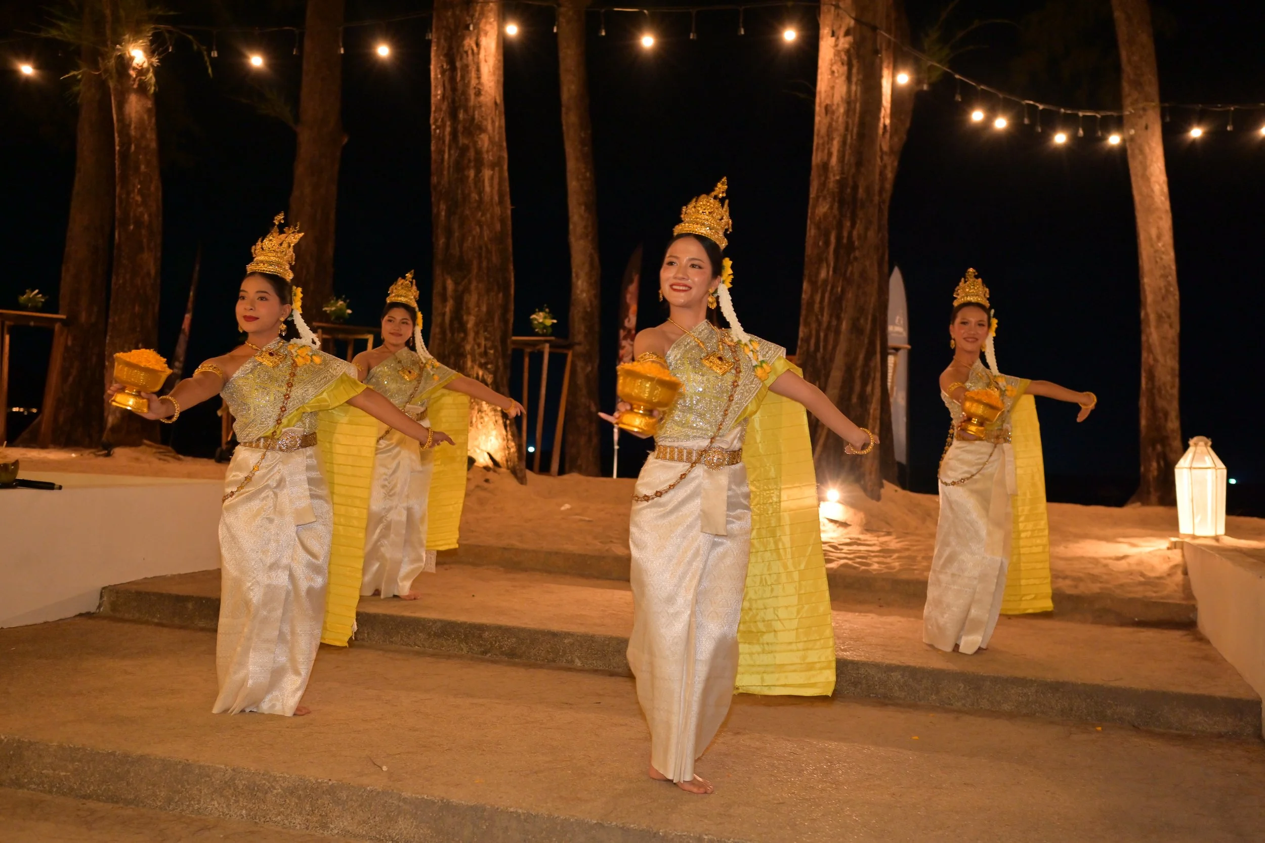 Four women in traditional Thai attire dancing at night outdoors, illuminated by string lights and standing on sandy ground among large trees.
