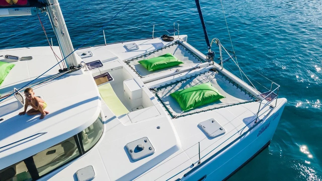 A white sailing yacht with green pillows on the deck, a boy sitting on the roof, and the boat floating on blue water.