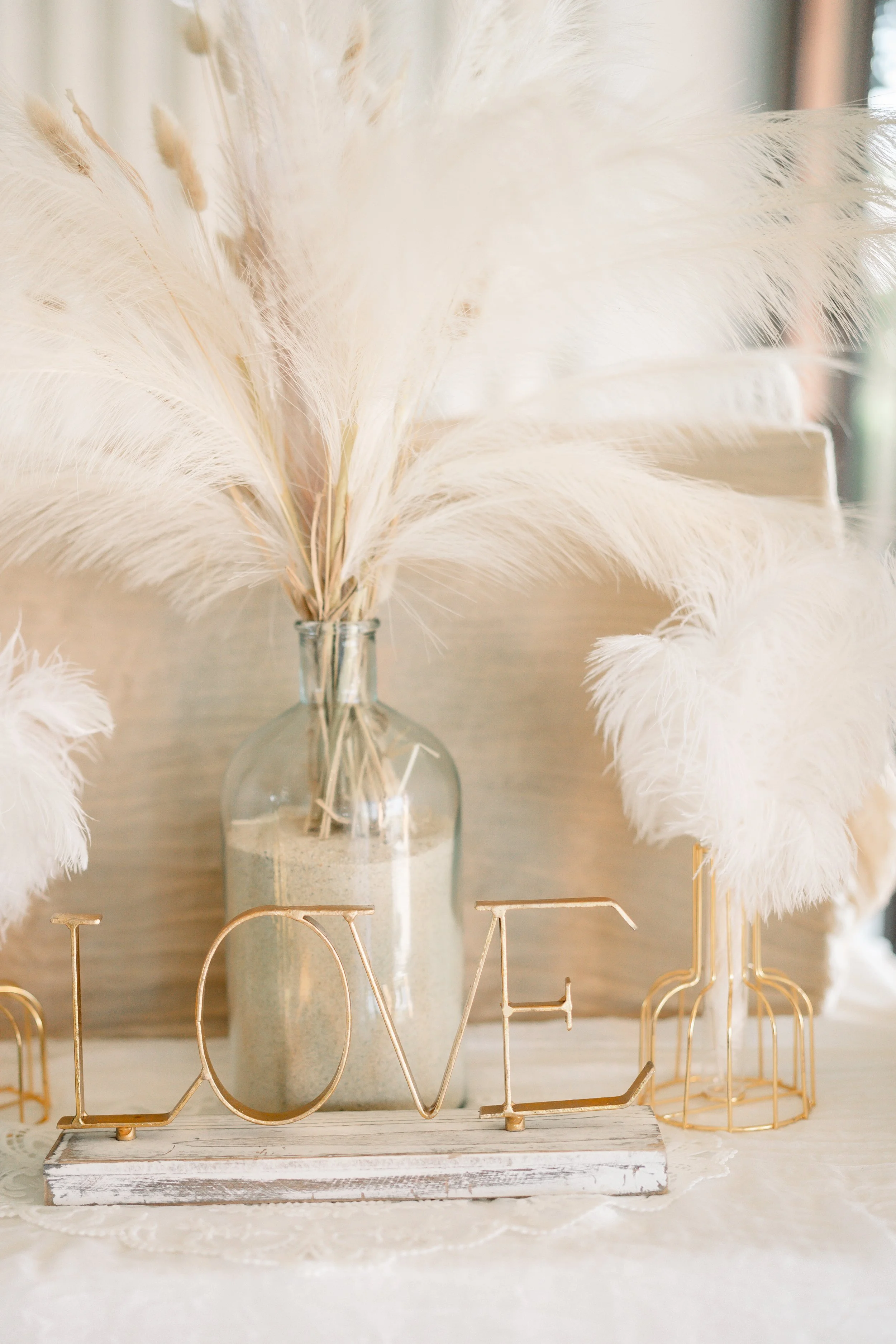 Decorative pampas grass arrangement in a glass vase with sand, placed behind gold 'LOVE' and 'DREAM' word signs on a white distressed wood surface, with a blurred background.