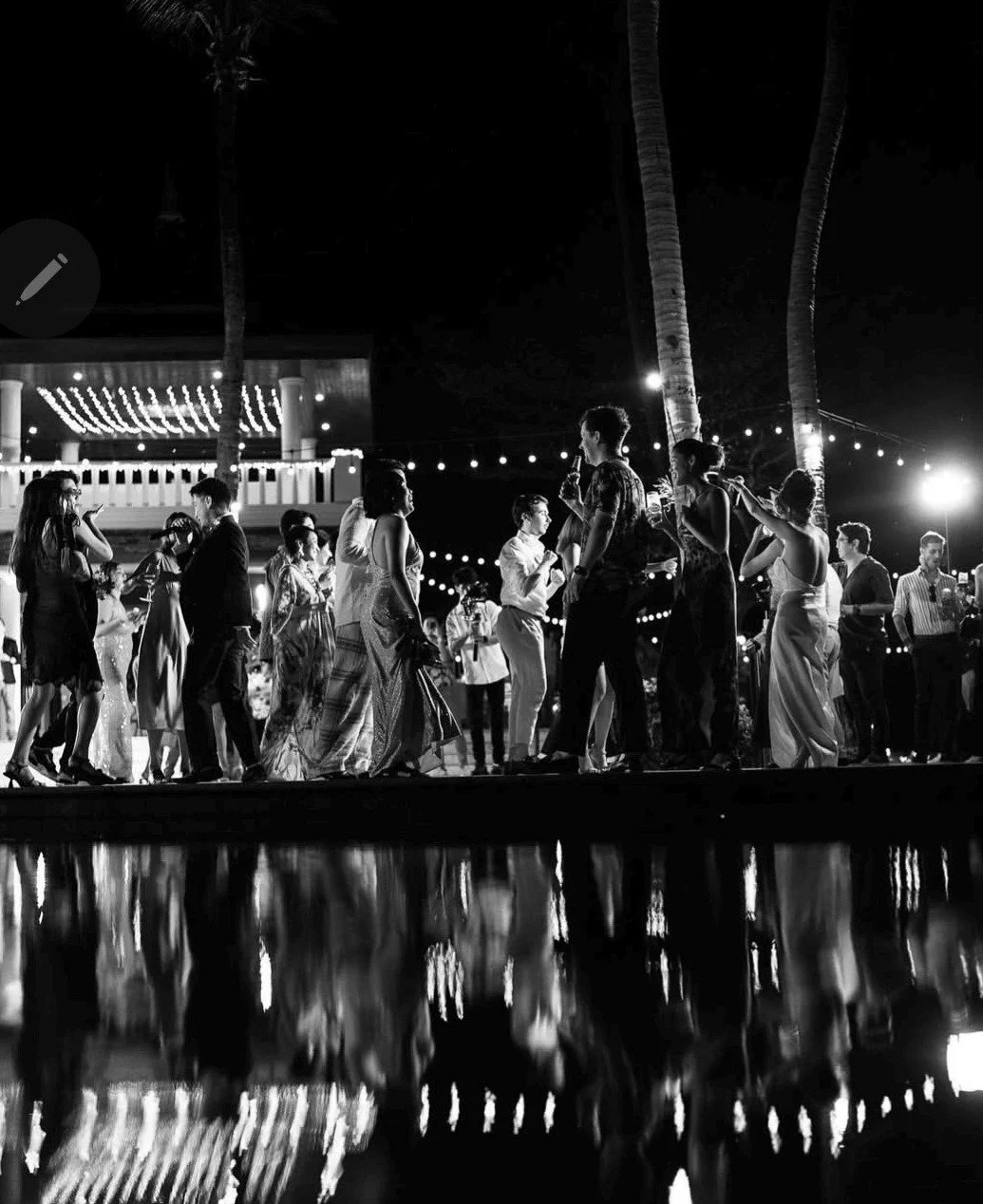 People dancing and socializing at an outdoor nighttime event with string lights and palm trees, reflected in water.