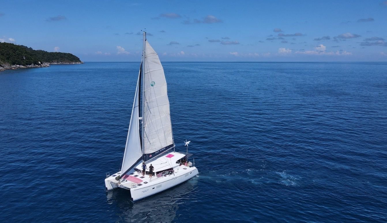A white sailboat with black and pink accents sailing on calm blue ocean water near a green hilly coastline under a partly cloudy sky.