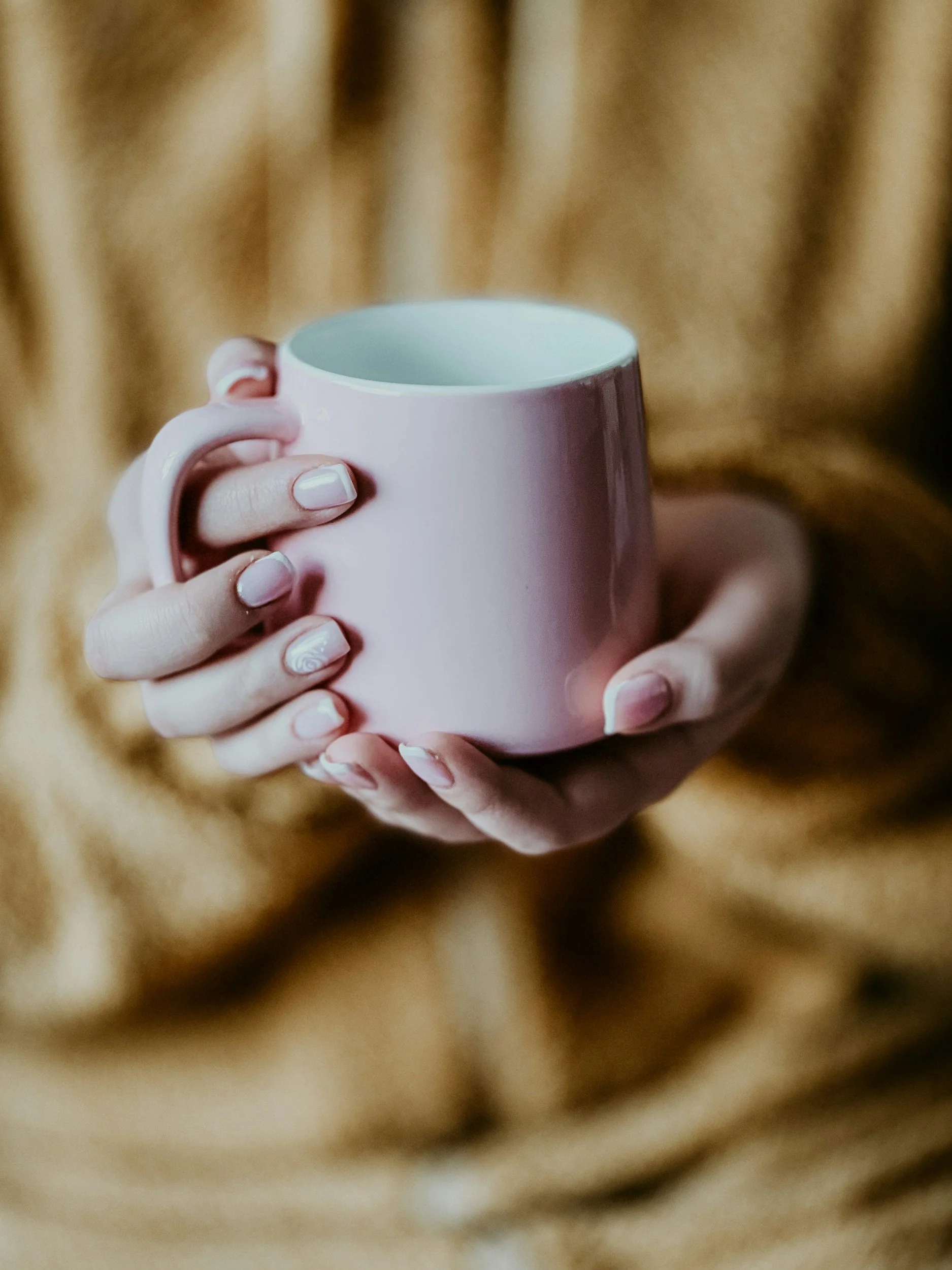 Close-up of a person holding a pink ceramic mug with both hands, focus on hands and mug, with a brown fur background.