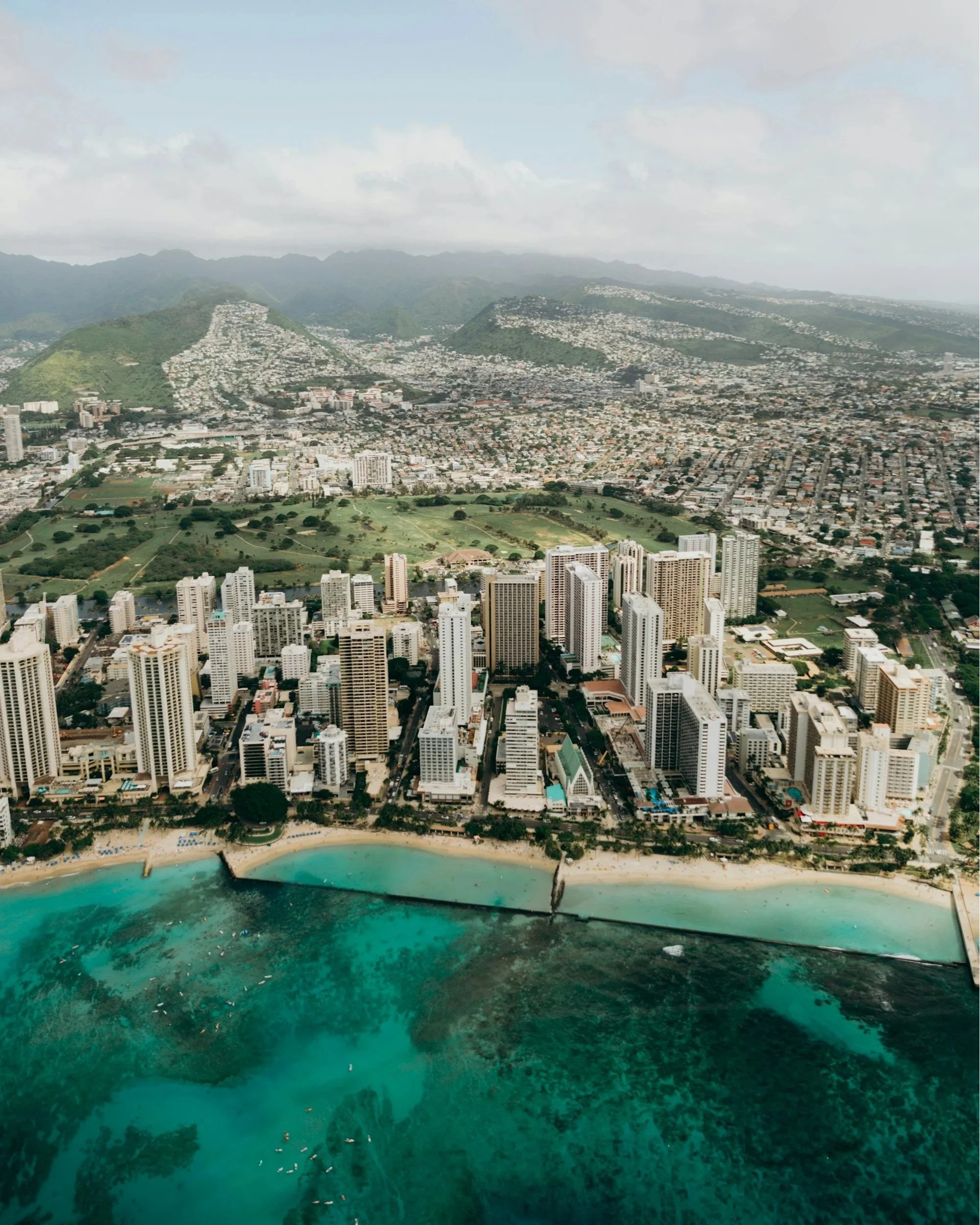 Aerial view of Honolulu with high-rise buildings along the coast and a turquoise ocean dotted with surfers, with green hills and mountains in the background.