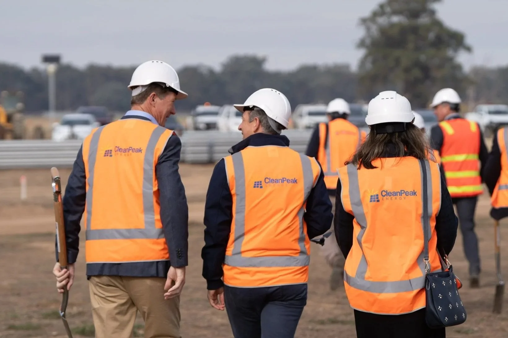 Group of CleanPeak Energy workers in high-vis safety vests and white hard hats walking on a solar farm construction site, with vehicles and trees in the background.