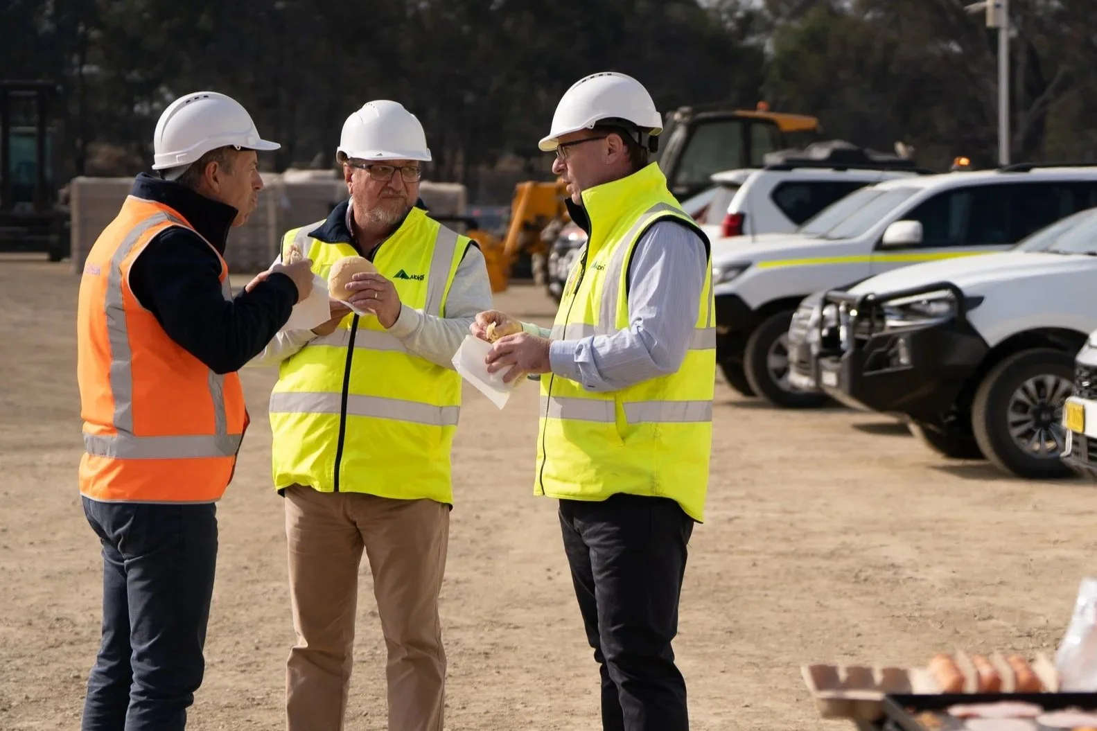Three men in safety vests and hard hats standing outdoors, eating sandwiches during a break. Background shows parked cars and construction machinery.