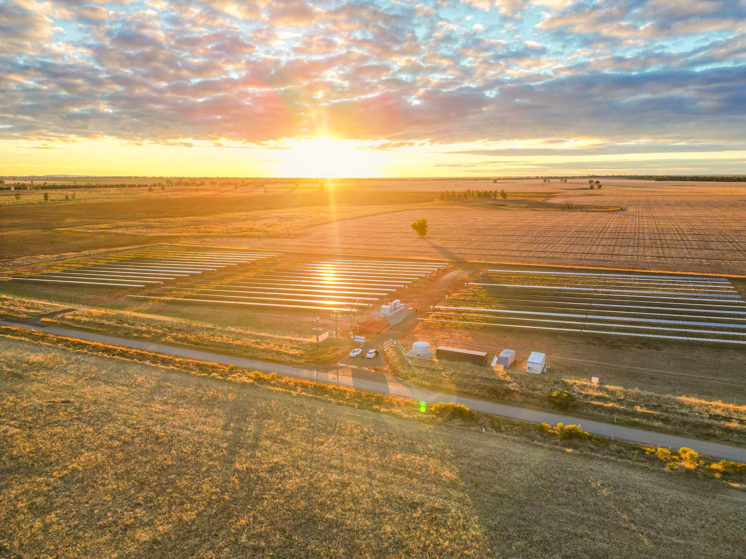 Sunset over a CleanPeak Energy solar farm in a field, showing solar panels, trees, and a partly cloudy sky.