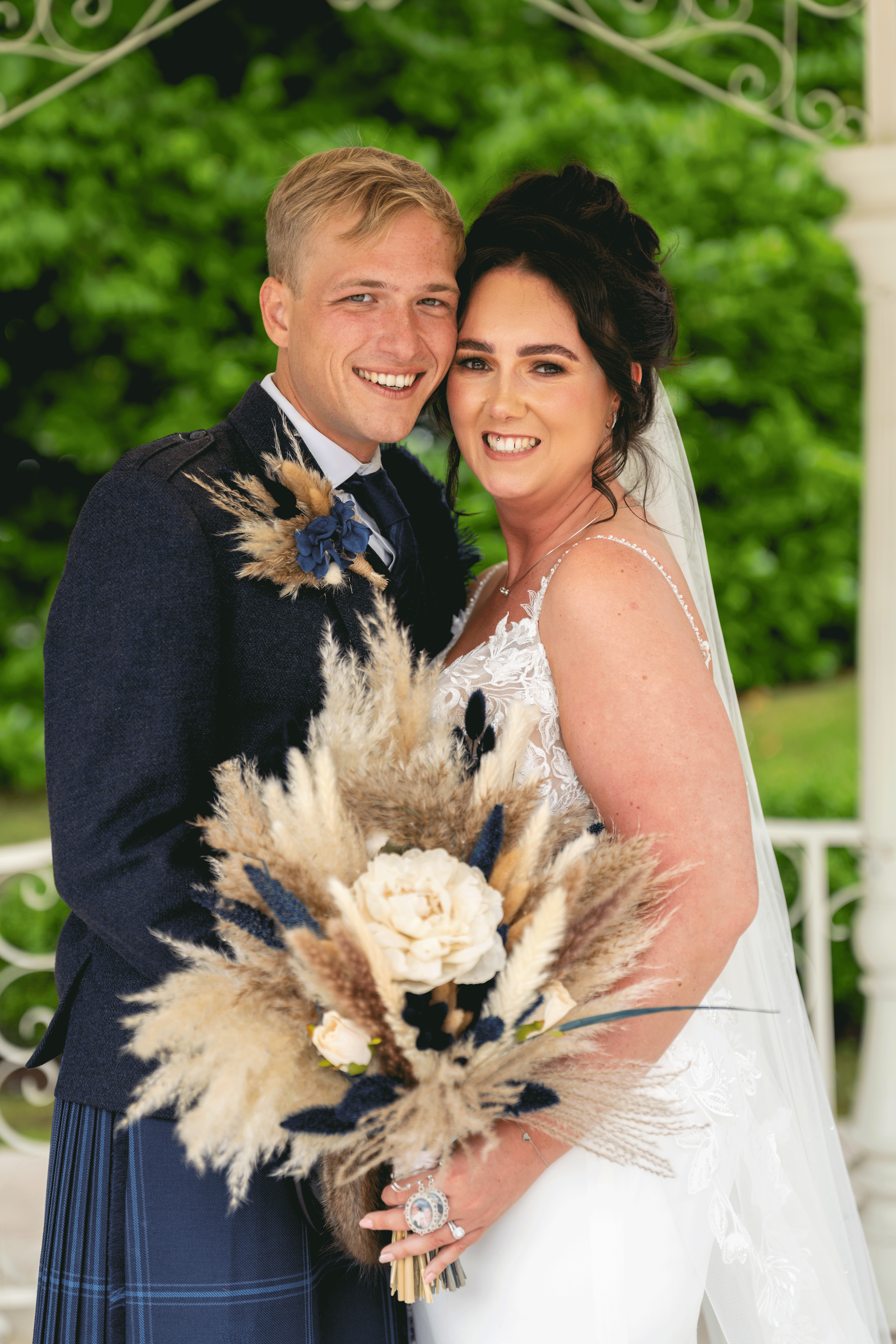 A bride and groom smiling and posing for a photo outdoors, with green foliage in the background. The bride is wearing a white lace wedding dress and holding a bouquet of dried flowers, and the groom is in a dark suit with a boutonniere.