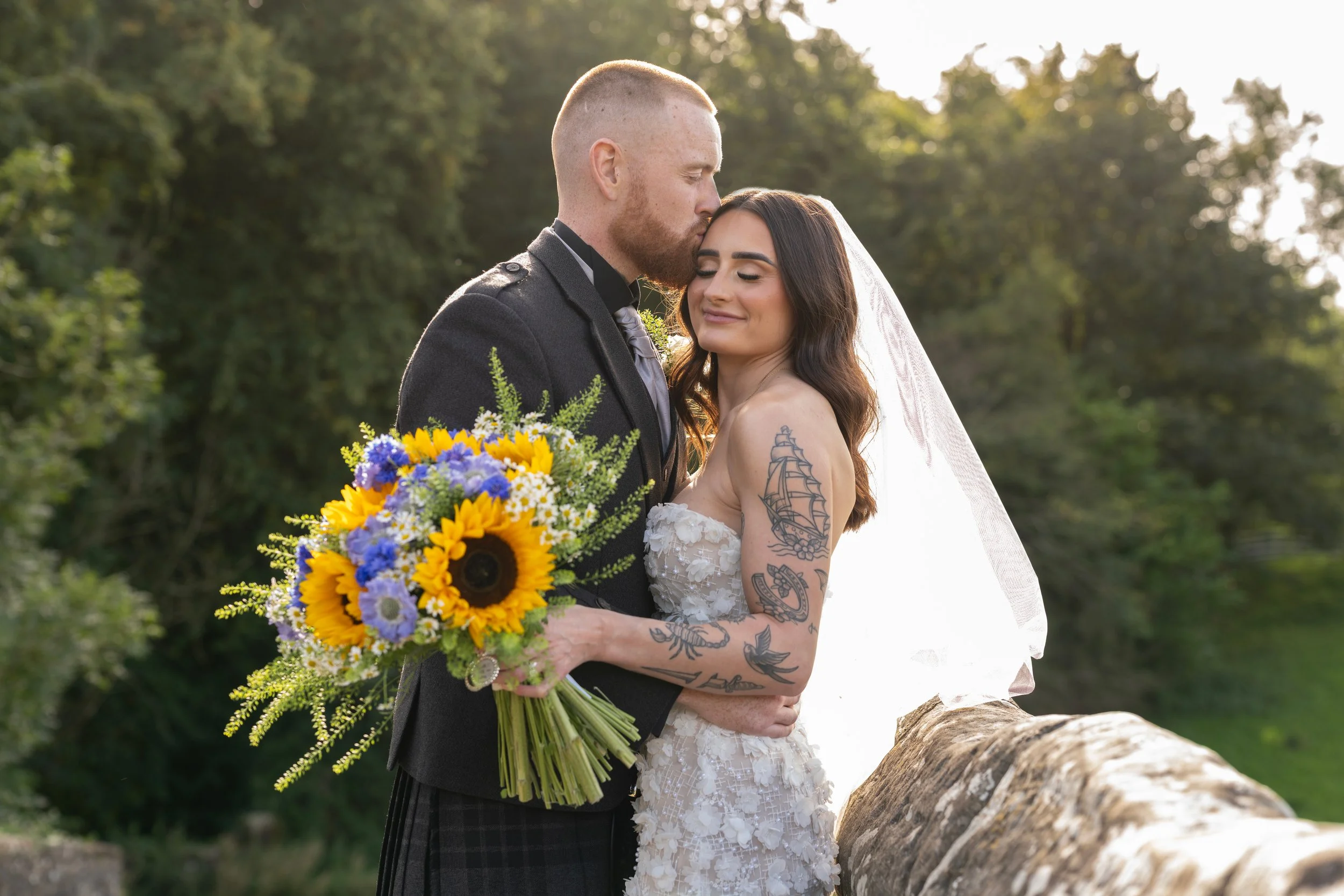 A newlywed couple embracing outdoors during sunset, with the bride smiling and holding a bouquet of white flowers. The groom has his eyes closed and is wearing a dark suit, while the bride is wearing a white wedding dress and veil.