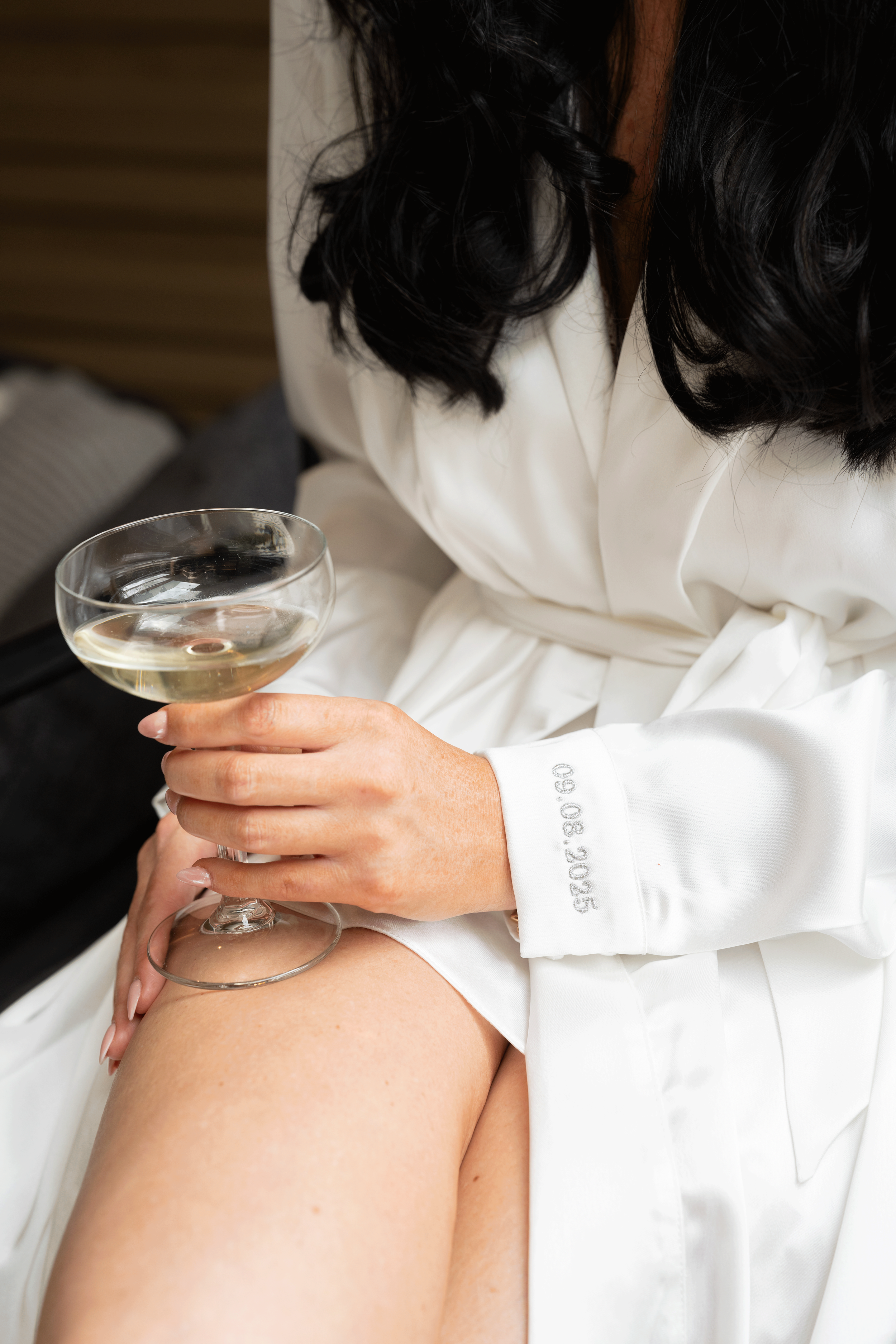 A Bride in a white satin robe with black hair holding a glass of white wine, sitting with a bare shoulder exposed.