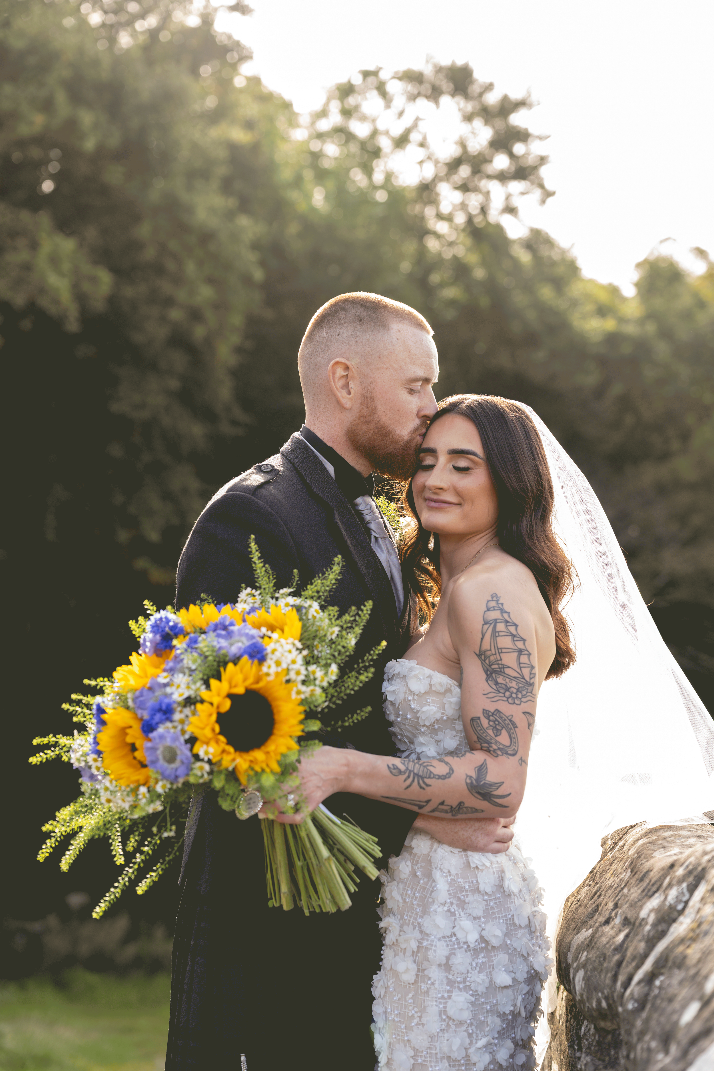 A newlywed couple sharing a tender moment outdoors during their wedding celebration. The man, with a beard and short hair, is dressed in a dark suit, gently kissing the woman's forehead. The woman, with long dark hair and tattoos on her arms, is wearing a floral lace wedding dress and holding a vibrant bouquet of sunflowers, daisies, and purple flowers. They are standing next to a large log, with a background of lush green trees and bright sunlight.
