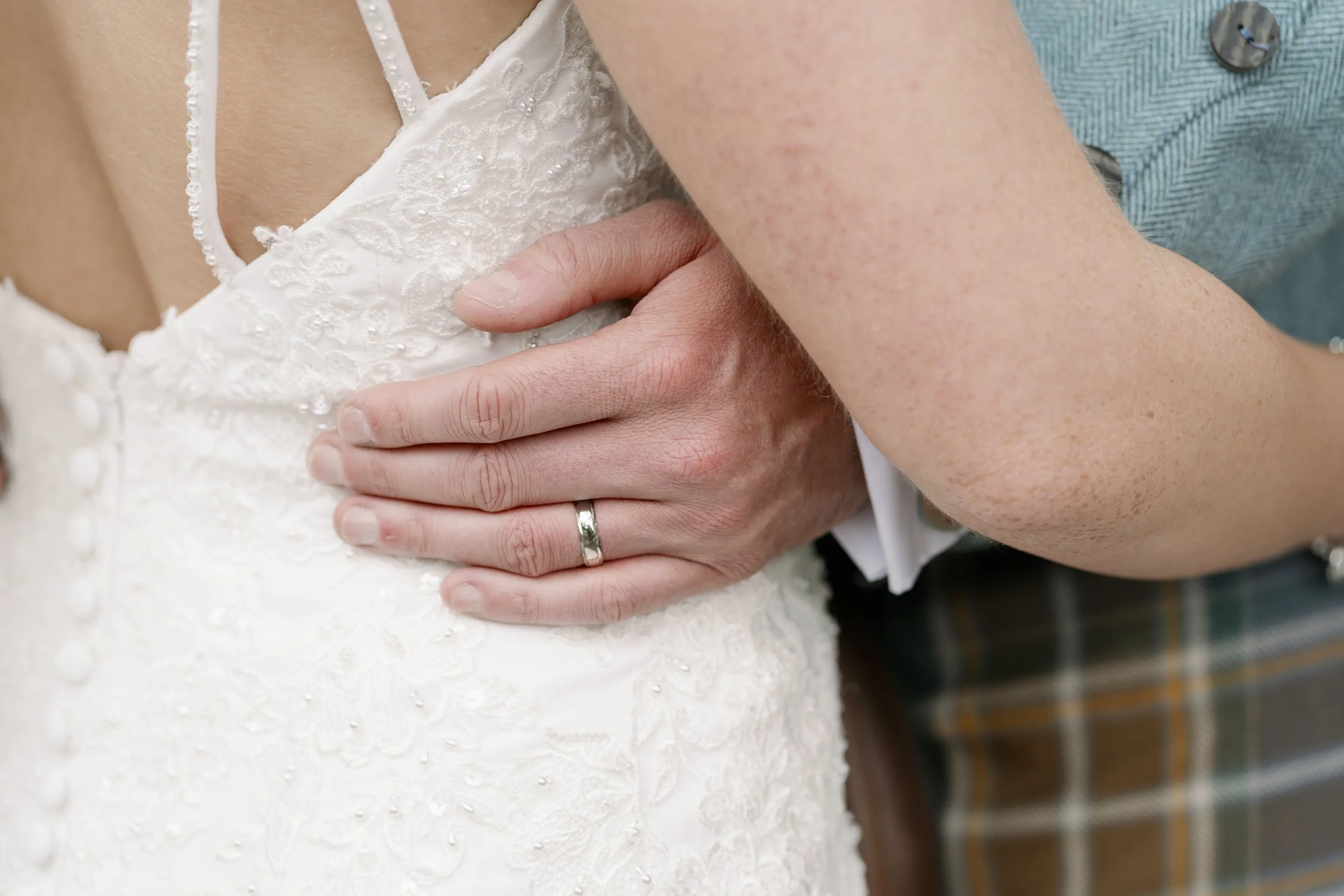 A close-up of a wedding couple's embrace, with one person wearing a white lace wedding dress and the other in a teal shirt, showing the man’s hand with a wedding ring resting on the woman's back during a wedding or special occasion.