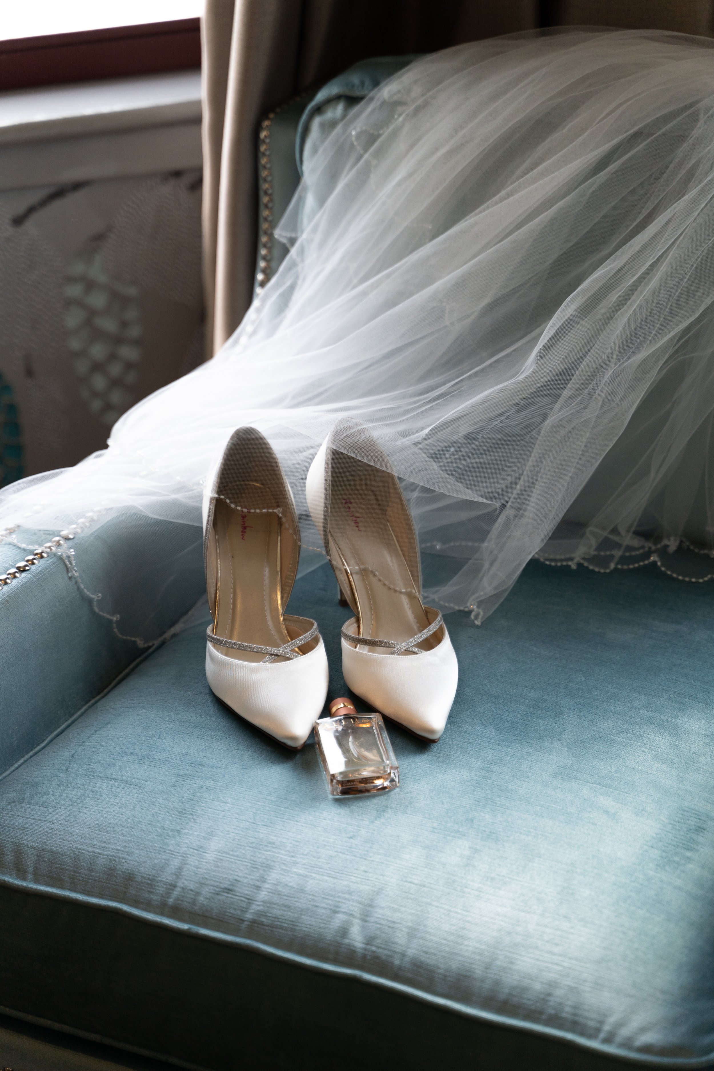 A pair of white bridal high-heeled shoes with gold accents placed on a teal fabric surface, with an engagement ring and clear acrylic block in front of them. Artistic wedding veil and jewelry are draped over the surface behind the shoes.