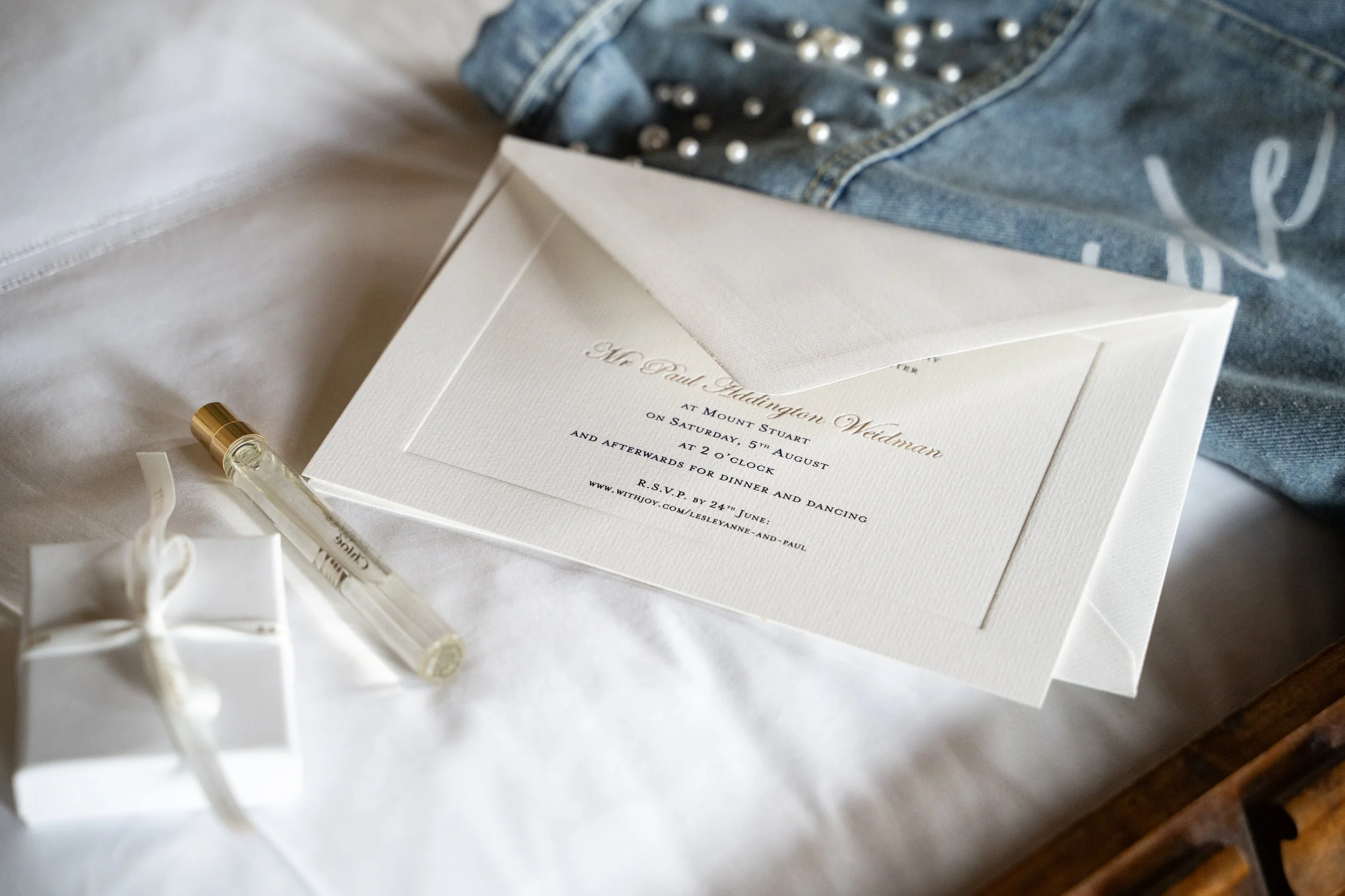 Wedding invitation card lying on a white surface, partially covered by a denim jacket with pearl embellishments. A white ribbon and a gold-trimmed pen are nearby.