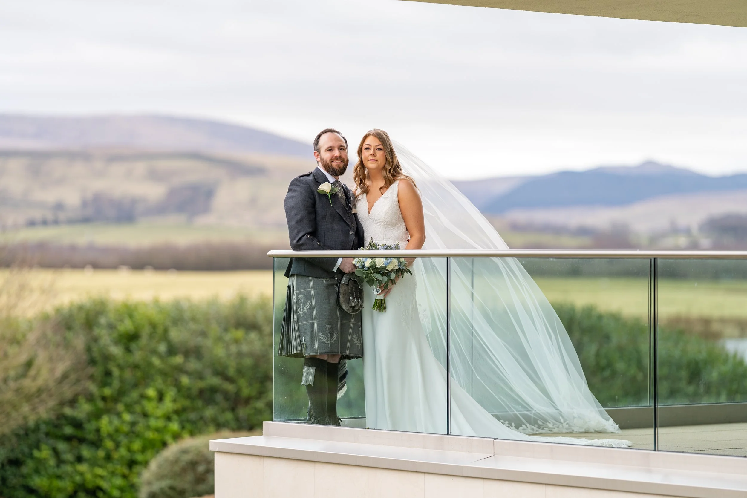 A bride and groom standing on a balcony with scenic mountains in the background. The groom is wearing traditional Scottish attire, including a tartan kilt, and the bride is dressed in a white wedding gown with a veil. They are holding a bouquet of flowers, and the groom has a beaming smile, while the bride has a more serious expression.