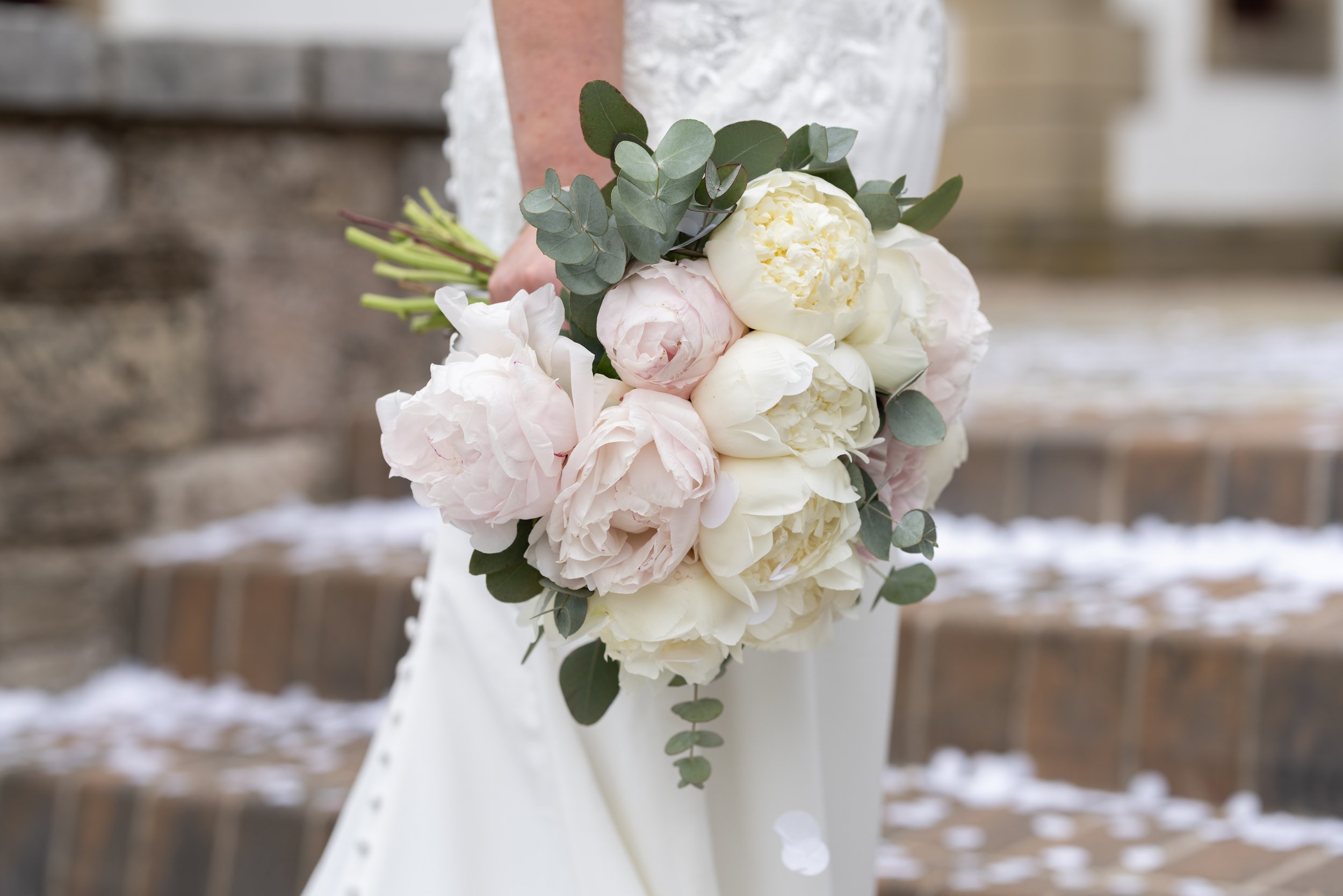 Bride in a white dress holding a bouquet of white and pink peonies, eucalyptus leaves, and green foliage with a brick and stone background.