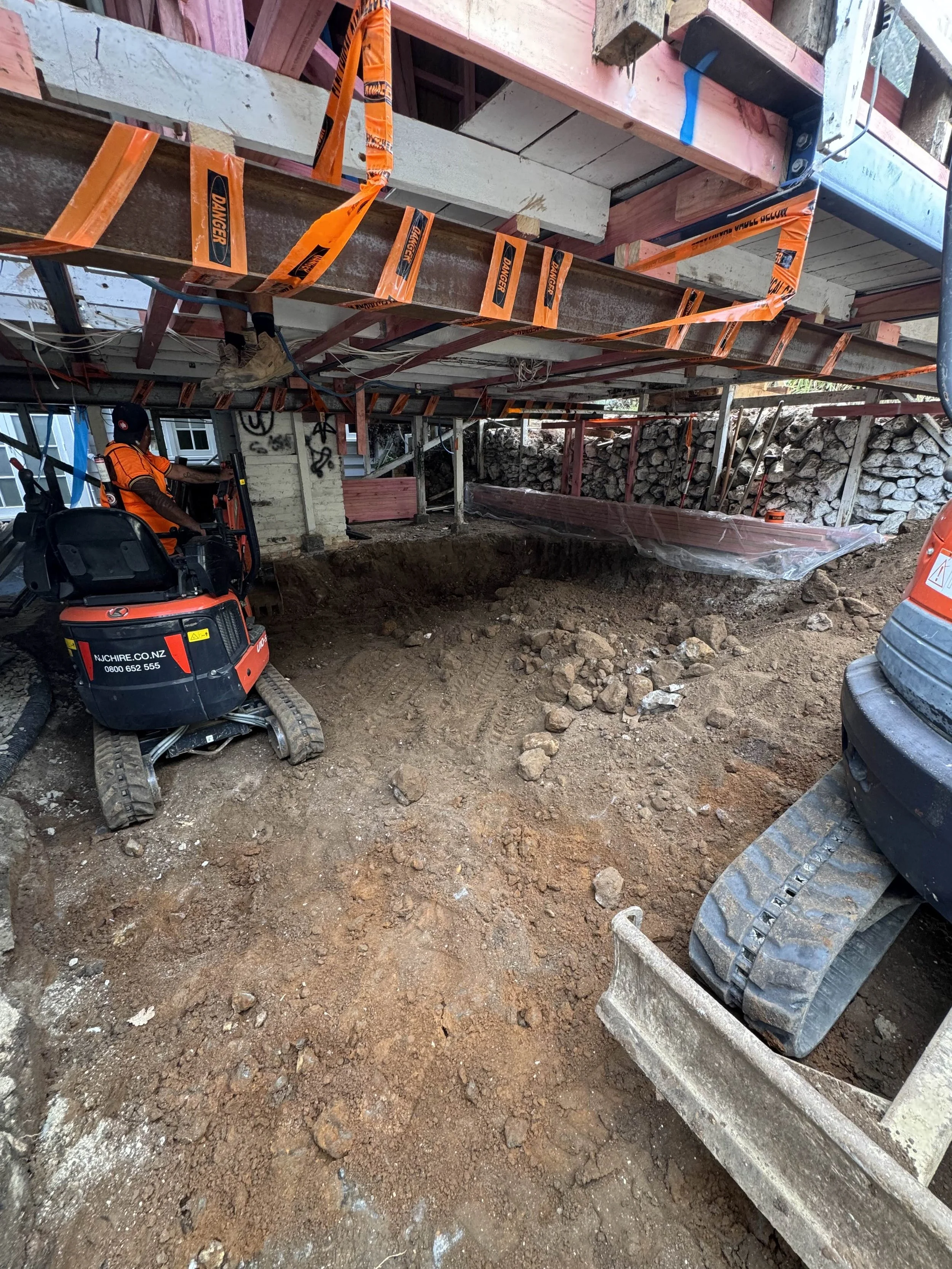 Construction site with exposed ground, construction materials, and workers. A worker operates a mini excavator, and there are wooden and metal structures supporting the building under construction. Orange warning tape is visible.