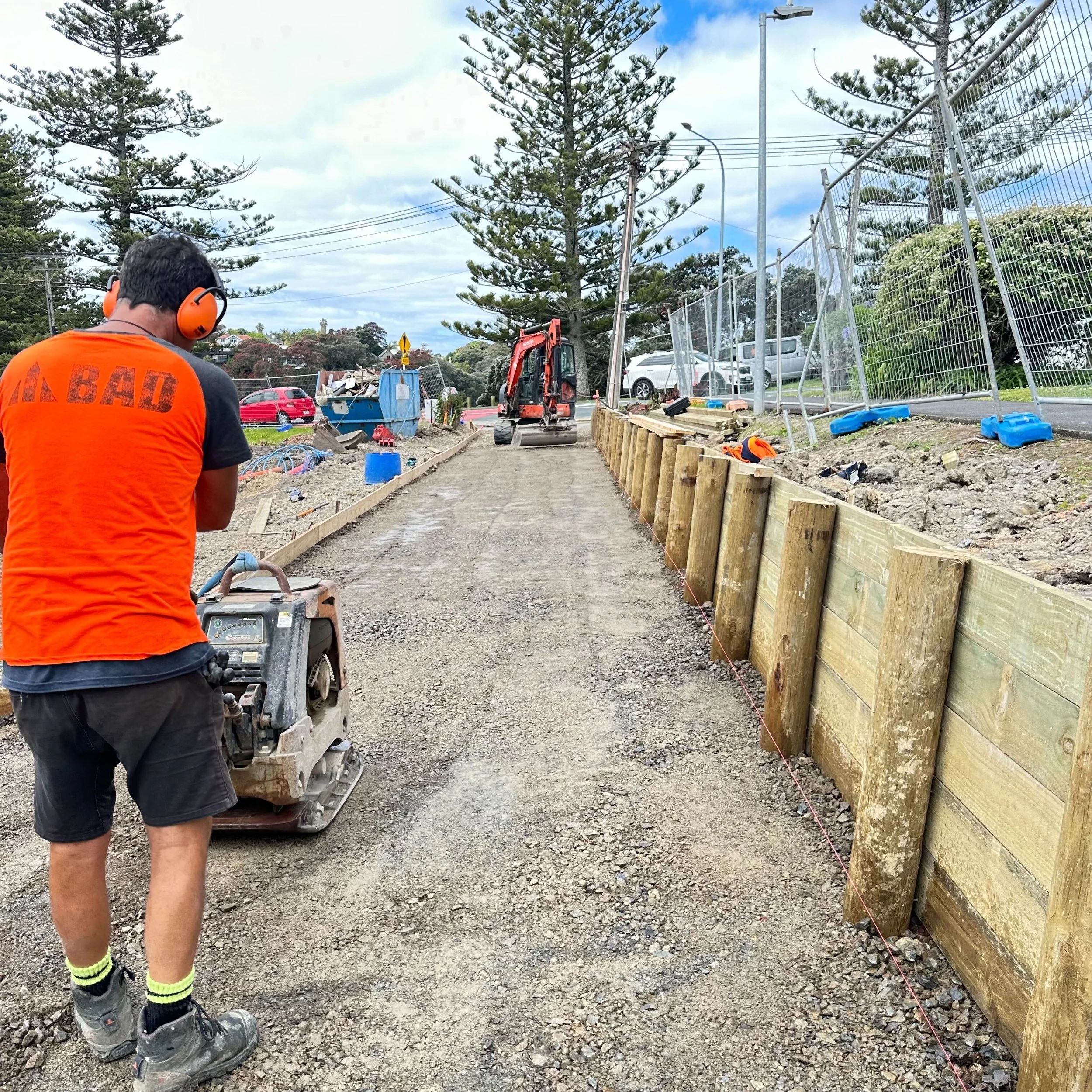 A construction worker wearing orange safety headphones, a bright orange safety vest, gray shorts, and work boots is operating a small utility vehicle on a gravel pathway at a construction site. In the background, there are tall pine trees, parked cars, construction equipment, and a partially assembled wooden barrier.