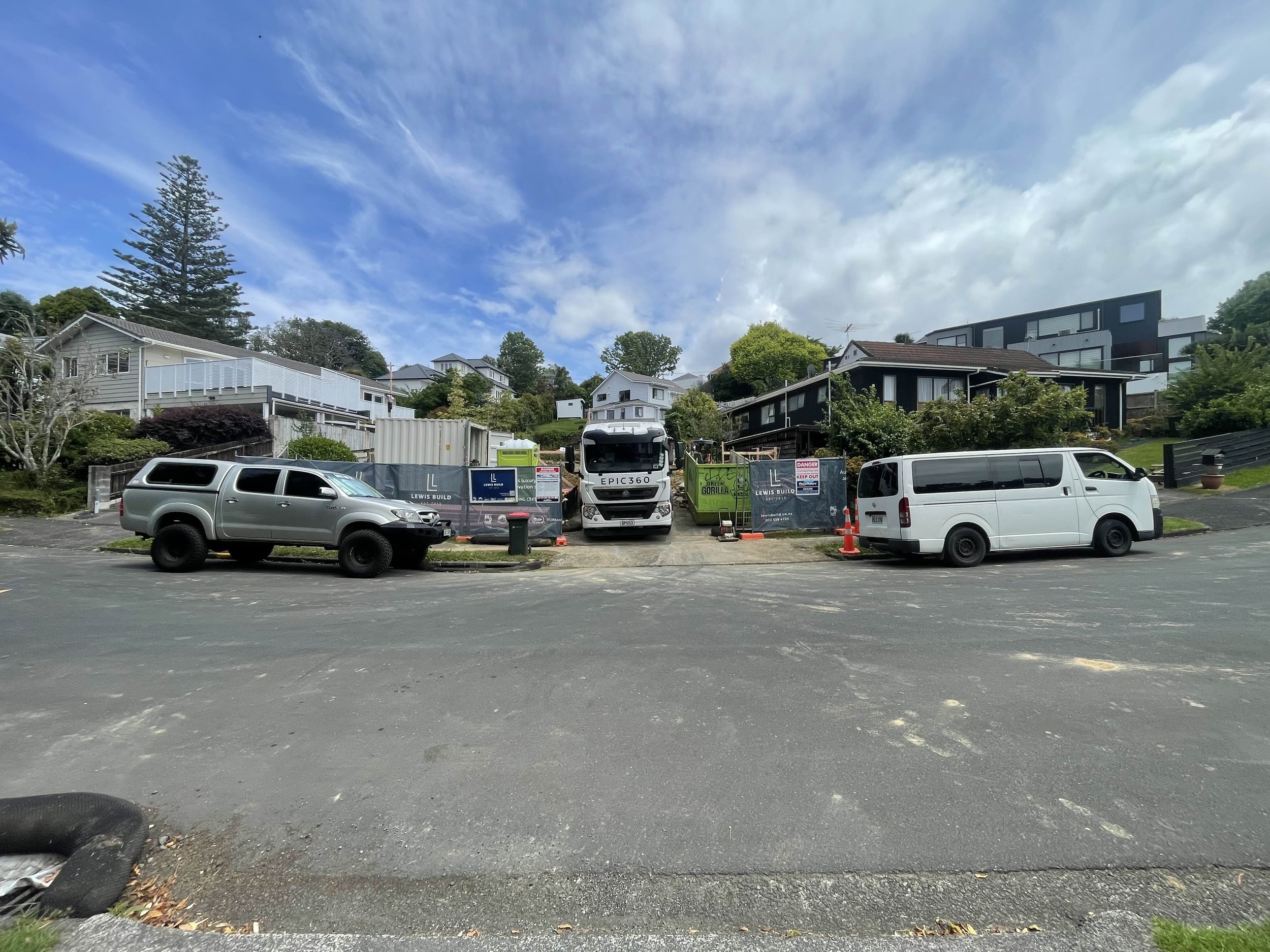 Residential street with parked cars in the foreground, houses on a hill in the background, and a partly cloudy sky overhead.