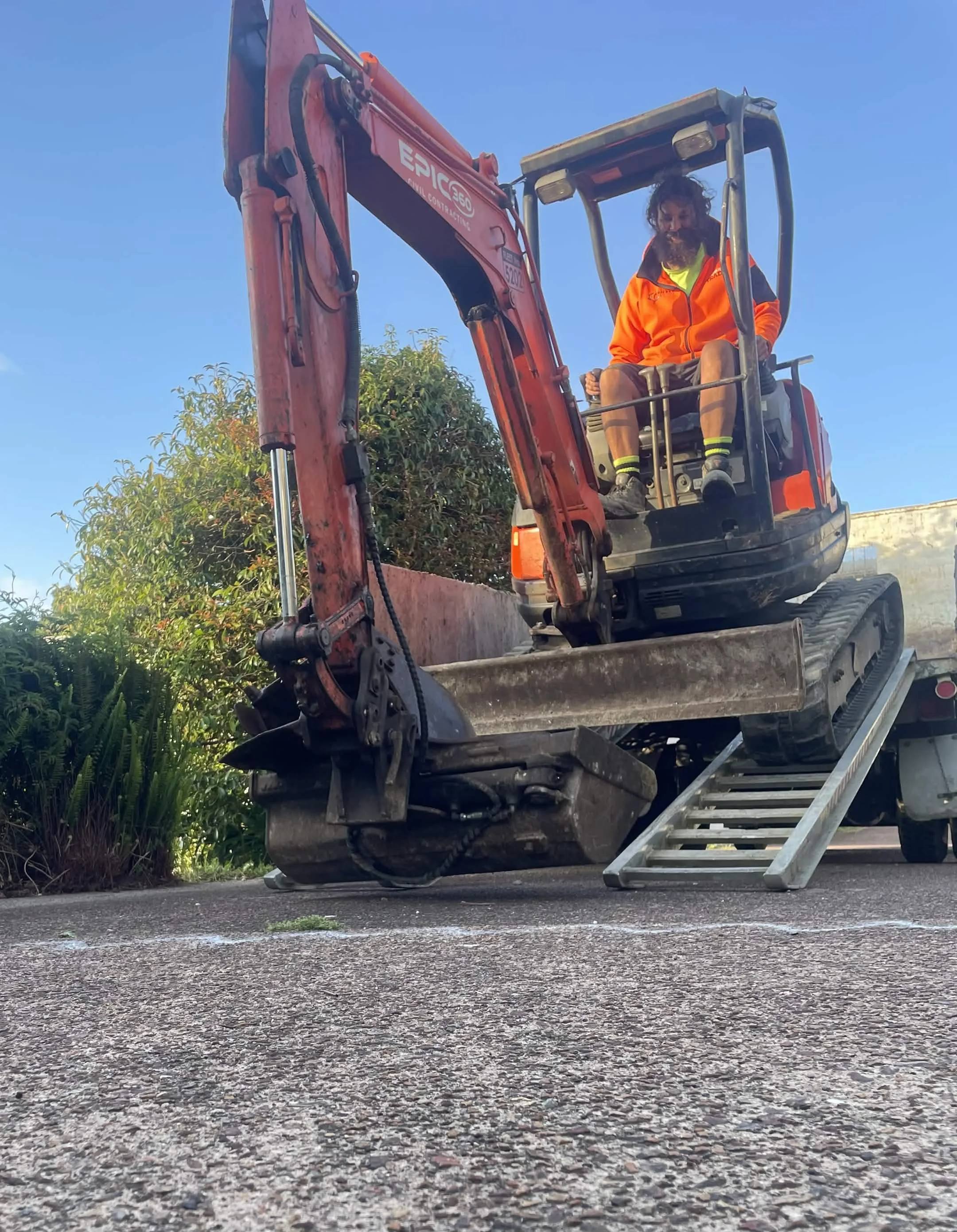 Person operating a small excavator with a ladder on the ground nearby. The person is wearing an orange jacket, shorts, and safety socks. The sky is clear and blue, and there are trees in the background.