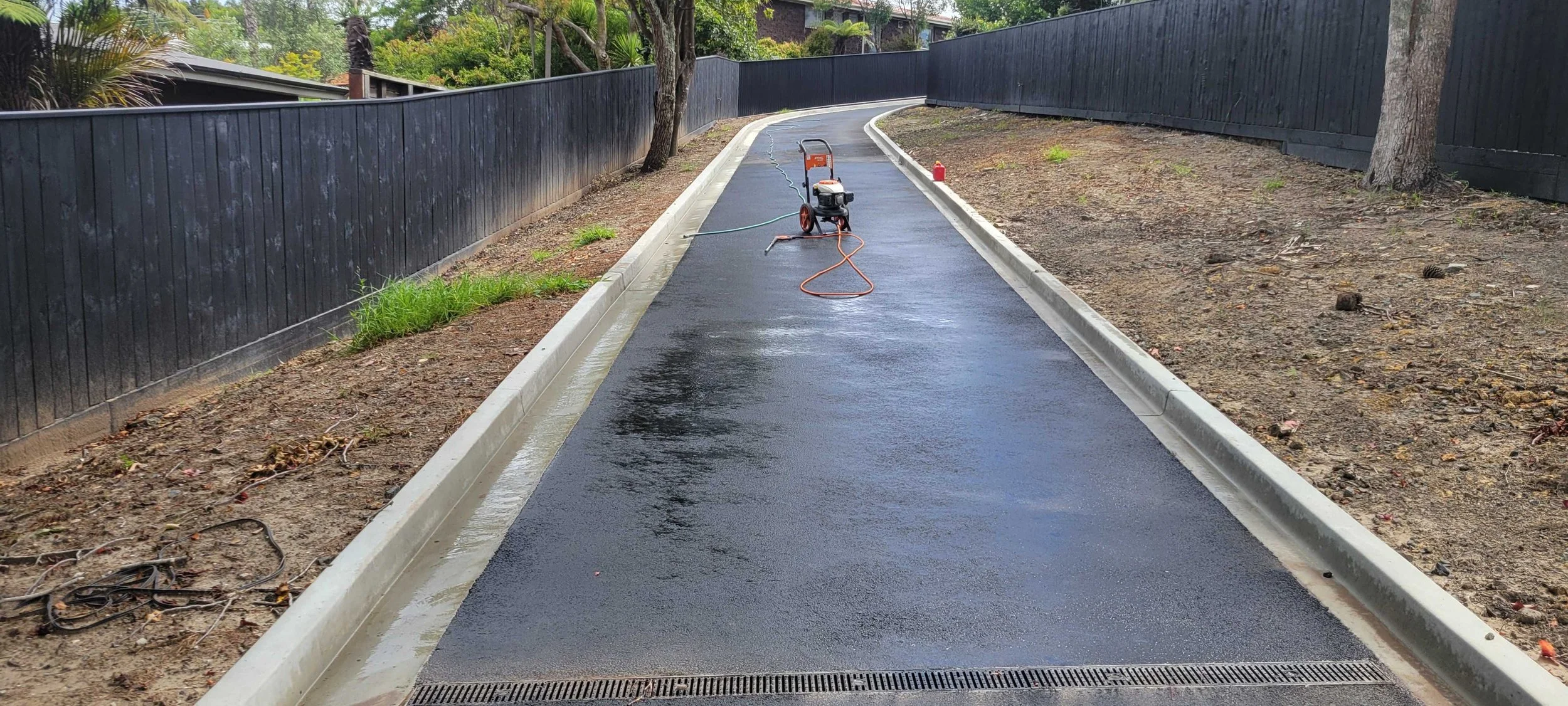 A freshly paved, wet asphalt sidewalk with a pressure washer, extension cord, and fire extinguisher placed on it, surrounded by a landscape with trees and a black wooden fence.