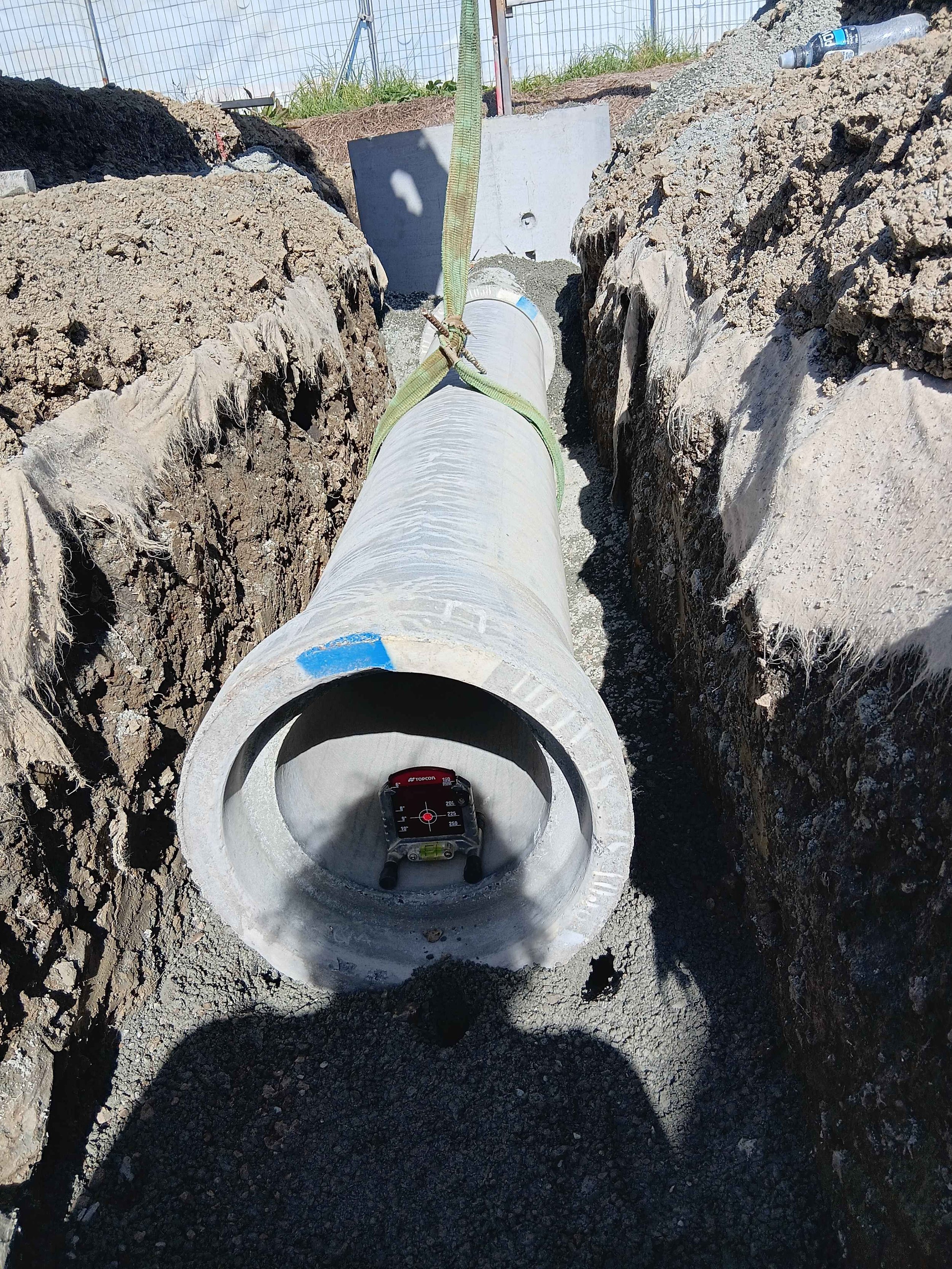 A large concrete pipe is installed underground, with a level and a laser distance meter placed inside it. The pipe is supported by a trench with dirt and gravel on either side, and a water bottle is lying on the ground nearby. A measuring tape is attached to the pipe, indicating construction or inspection work.