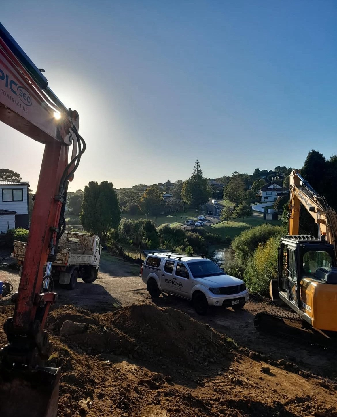 Construction site with excavators and a white vehicle marked 'Epic 360' on dirt, with houses, trees, and a pond in a suburban area in the background.