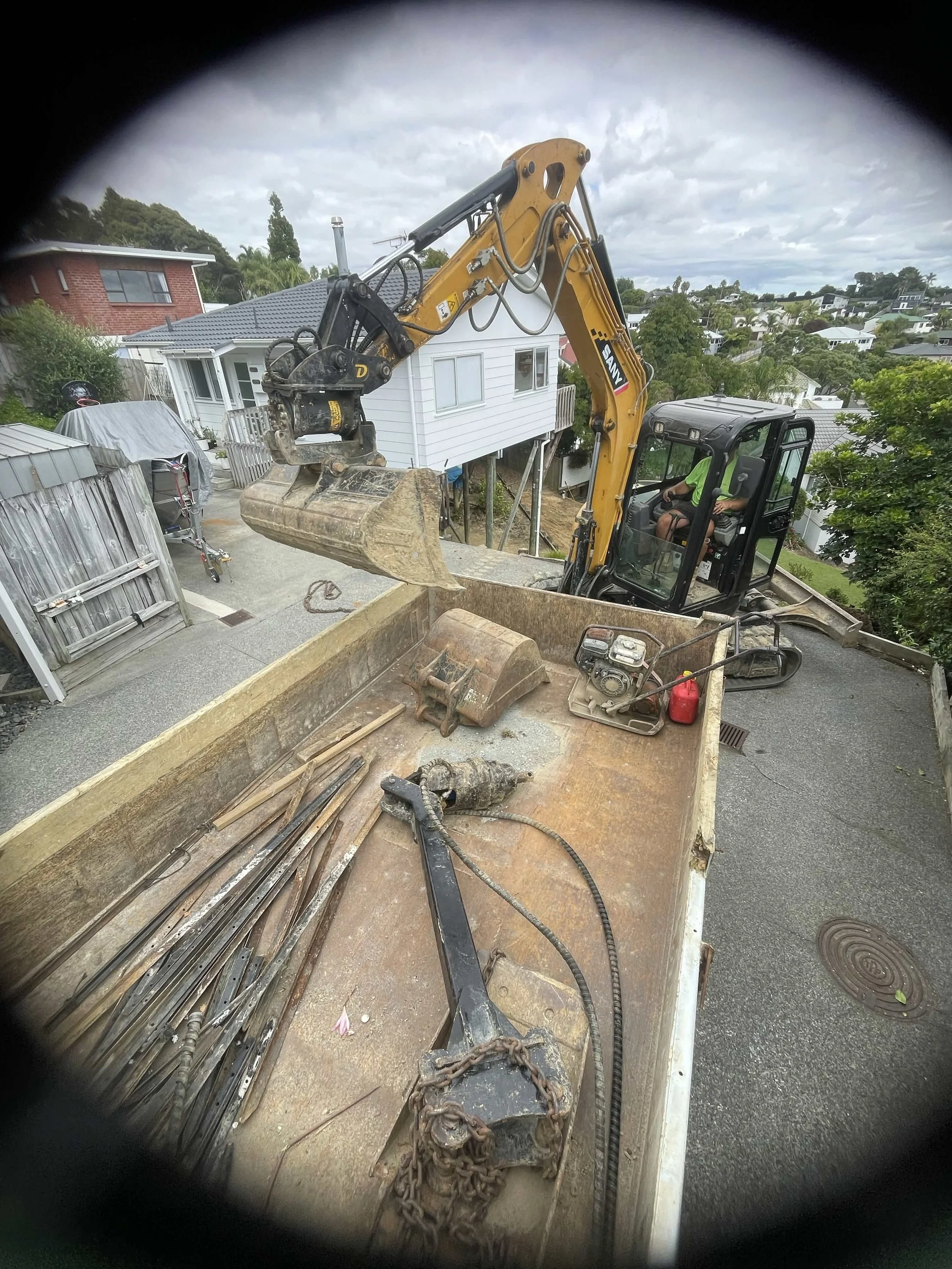 Excavator lifting a bucket filled with dirt inside a trailer at a construction site in a residential neighborhood.