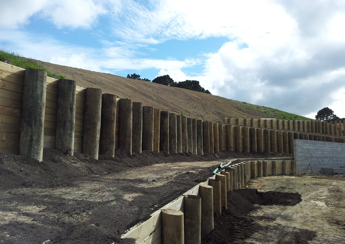 Construction site with wooden retaining wall on a hillside, partially completed, with a dirt path and some construction materials.