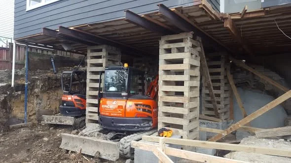 Construction site beneath a raised building with three support columns made of stacked concrete blocks. Two mini excavators are present, one orange and one black. Construction tools and materials are scattered around.