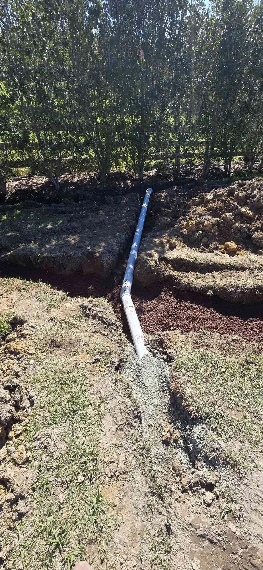 A long black and blue pipe extending from an underground hole in the ground toward a green bushy area, indicating a pipe installation or repair work.