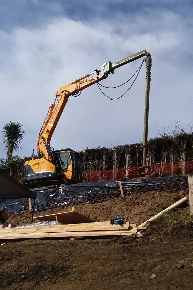 Construction site with a yellow Komatsu excavator lifting a steel rebar cage, workers in safety gear, and trees and houses in the background.