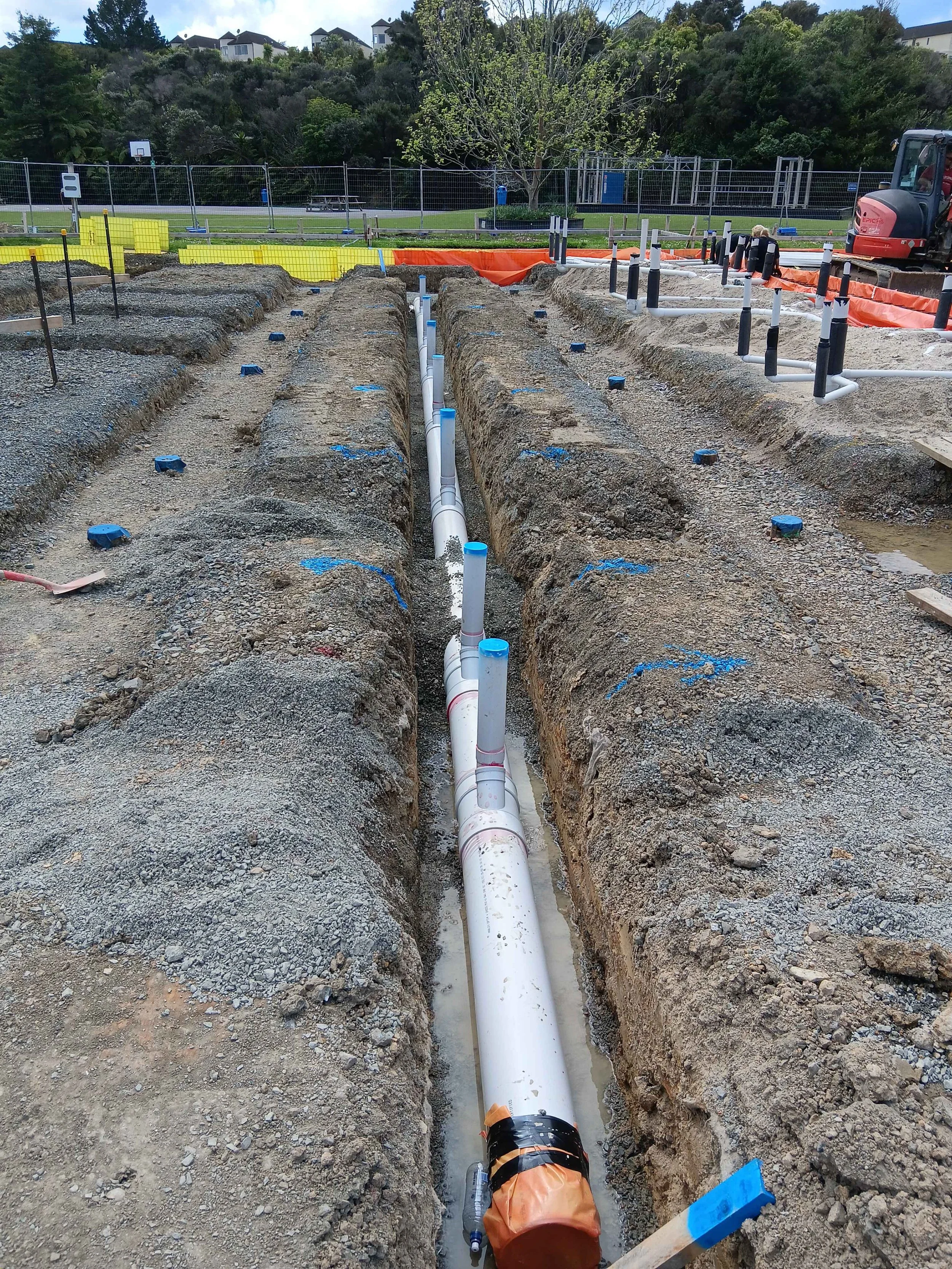 Construction site with large pipe in excavated trench, PVC piping with black and white markings, surrounded by dirt and gravel, orange and black tape on pipe, construction equipment and barriers in background.