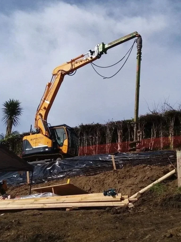 Construction site with an excavator pouring concrete into a vertical mold, with wooden planks and construction materials on the ground.