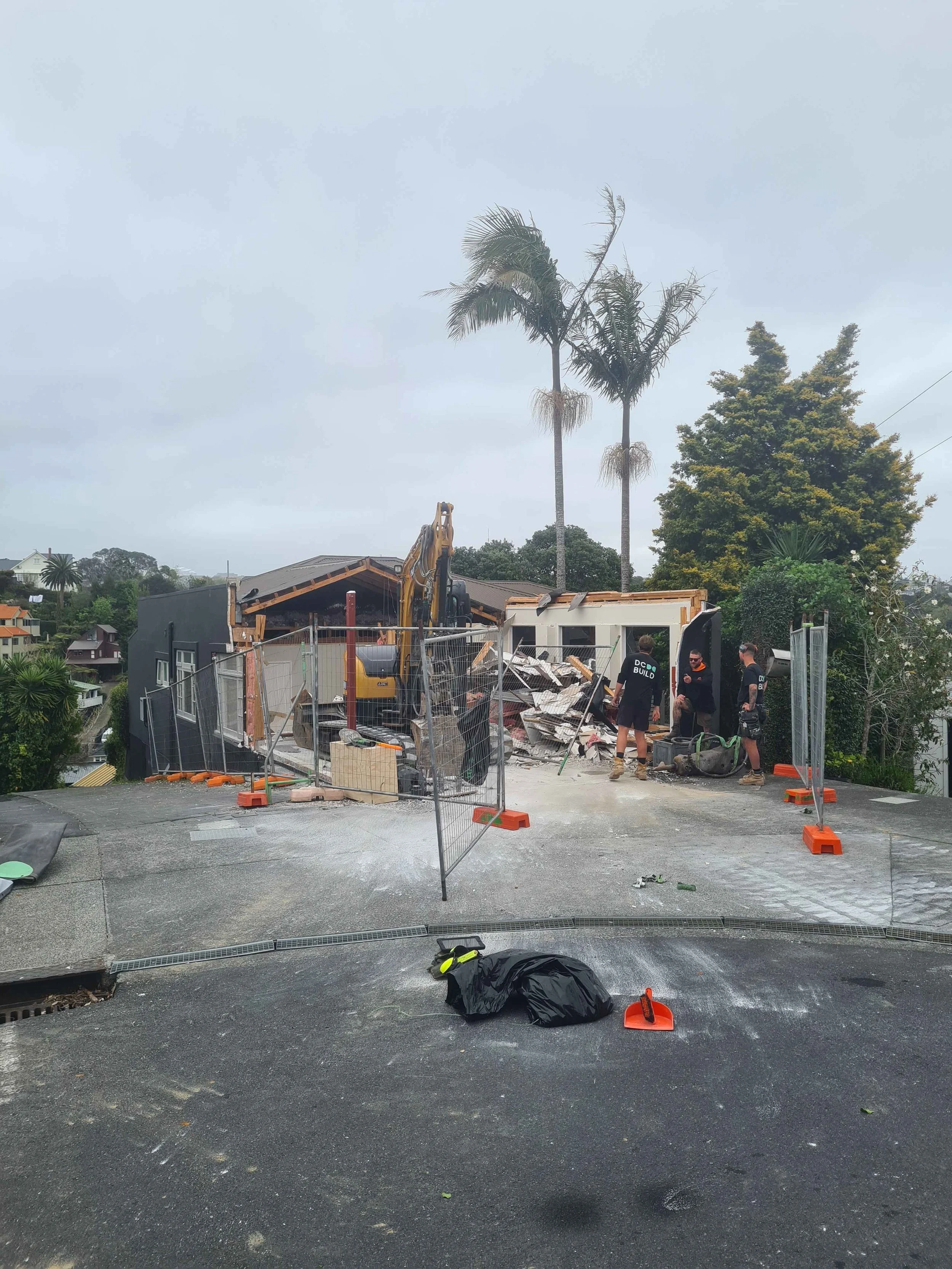 View of a construction or demolition site on a residential rooftop, with debris, safety barriers, and a backhoe, under an overcast sky.