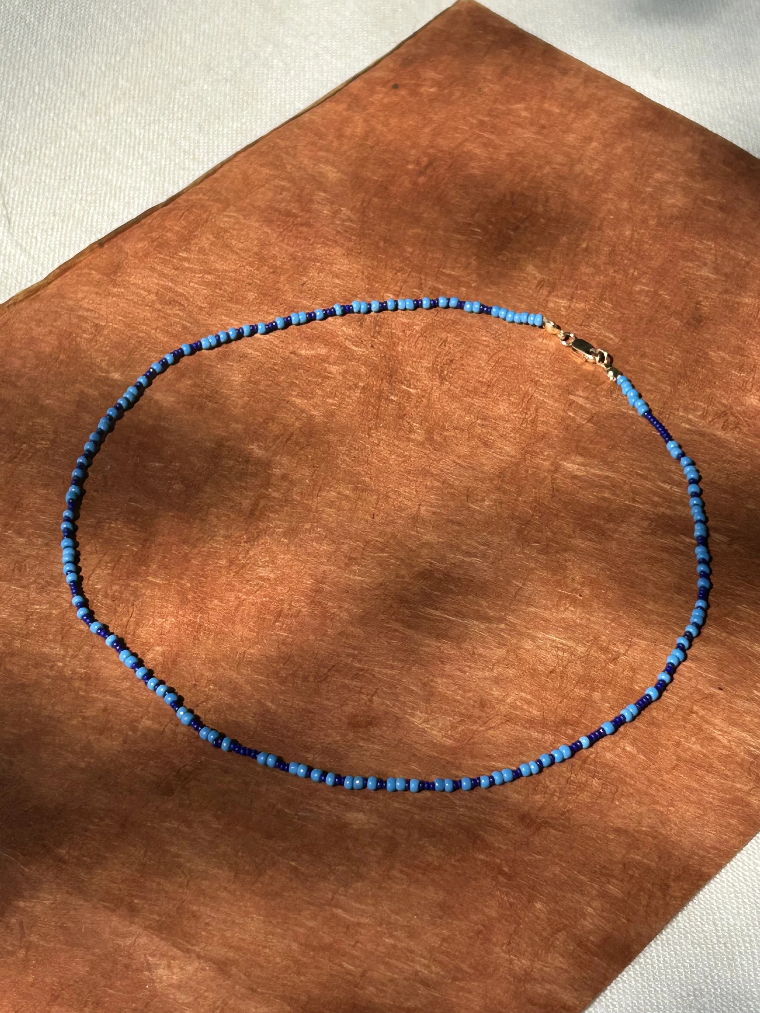 A beaded necklace with blue beads rests on a wooden surface with a white cloth nearby.