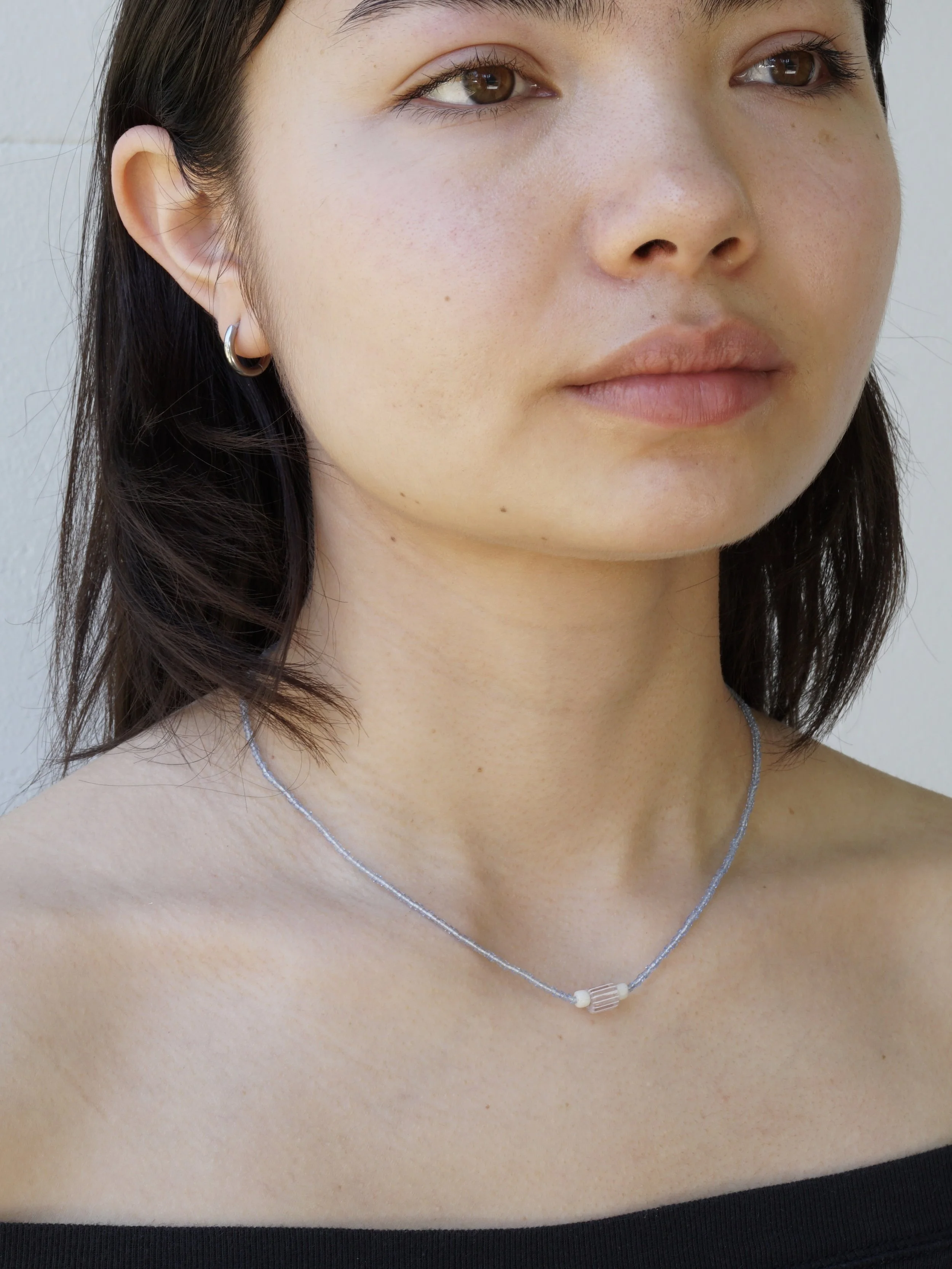 Close-up of a woman's face and upper chest, wearing a silver necklace and small hoop earrings, with dark hair and wearing a black off-shoulder top.
