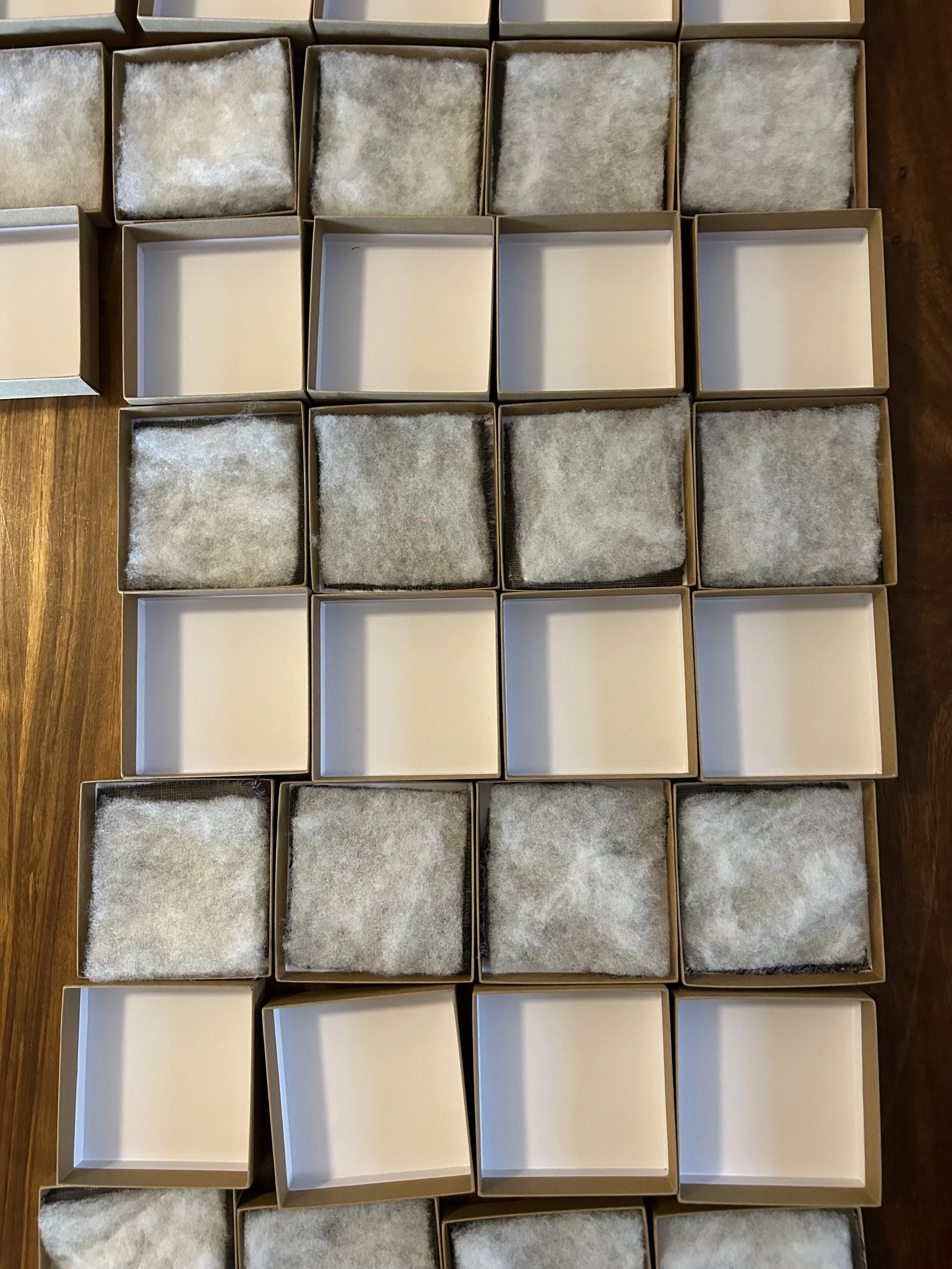 Multiple empty and partially filled cardboard boxes arranged in a grid on a wooden surface, some lined with cotton or similar material.