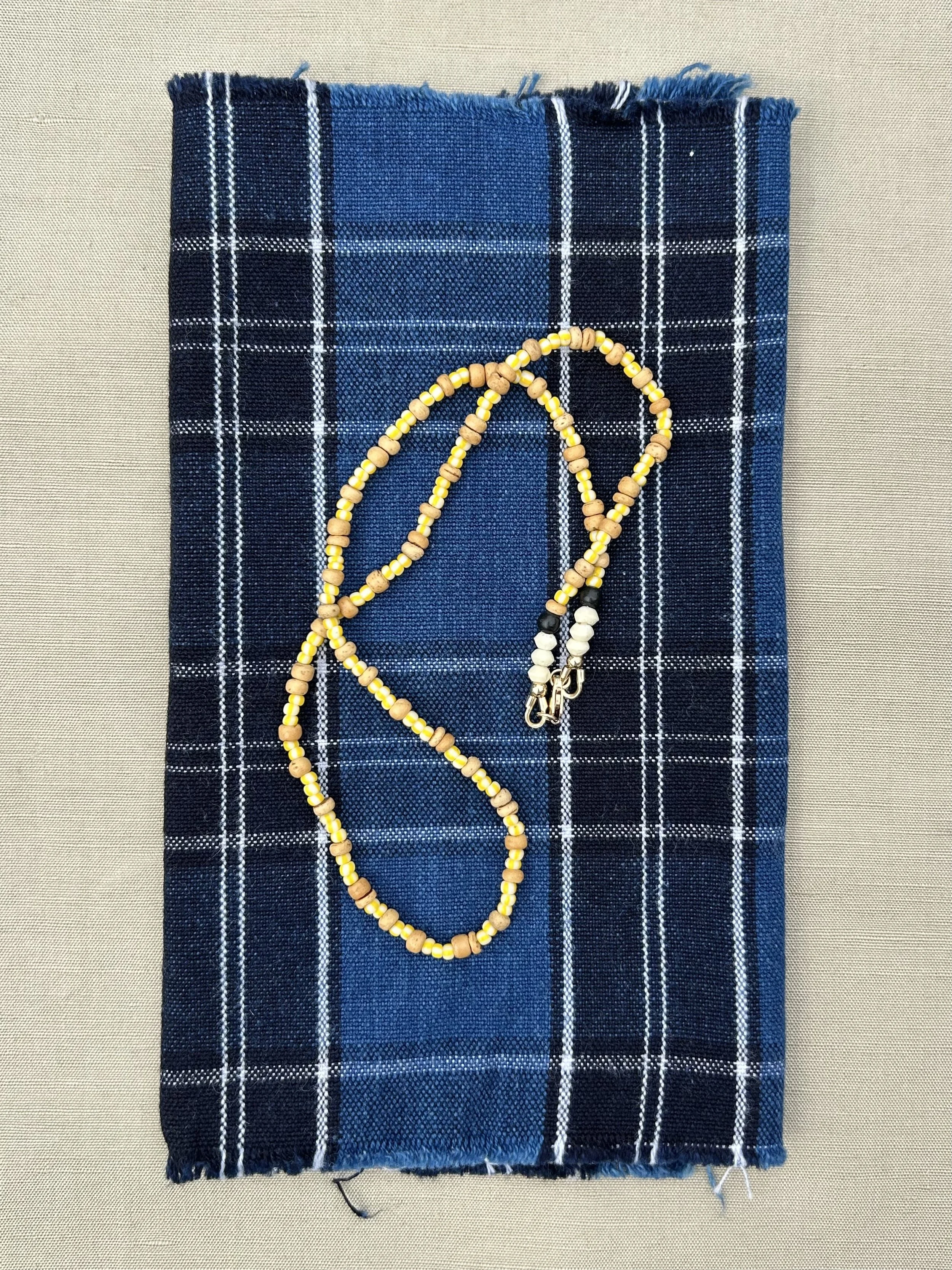 A vintage japanese blue plaid cloth napkin with fringed edges, placed on a recycled paper surface, with a beaded yellow, black, and white necklace arranged on top.