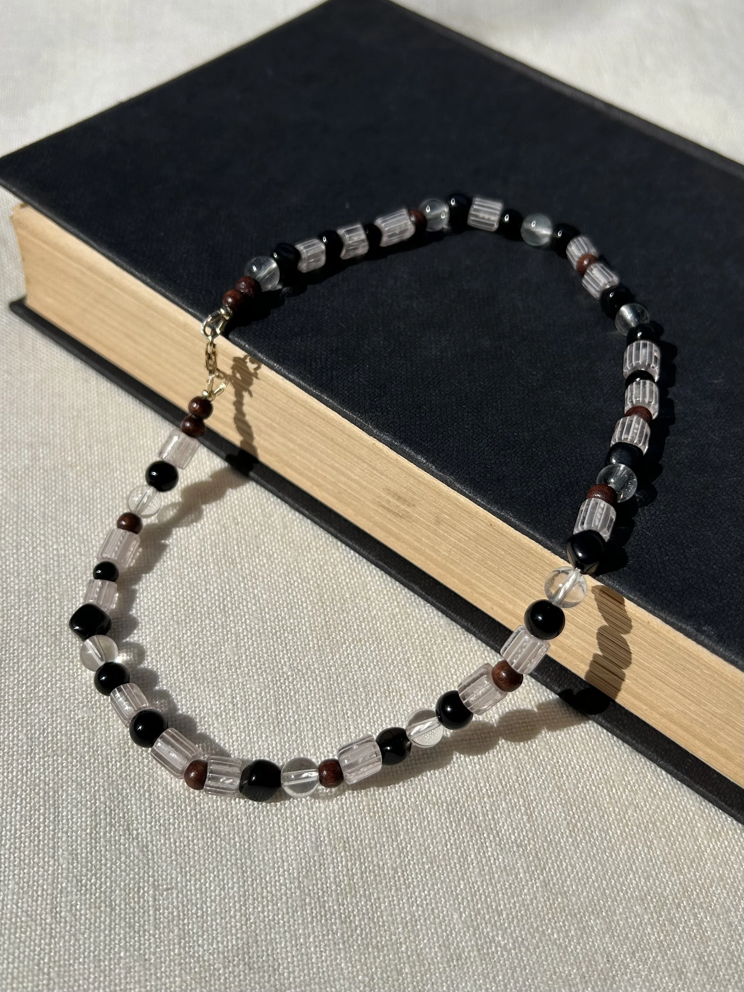 Beaded necklace with black, brown, and clear beads, draped over a closed black book with a textured cover, resting on a light-colored fabric surface.