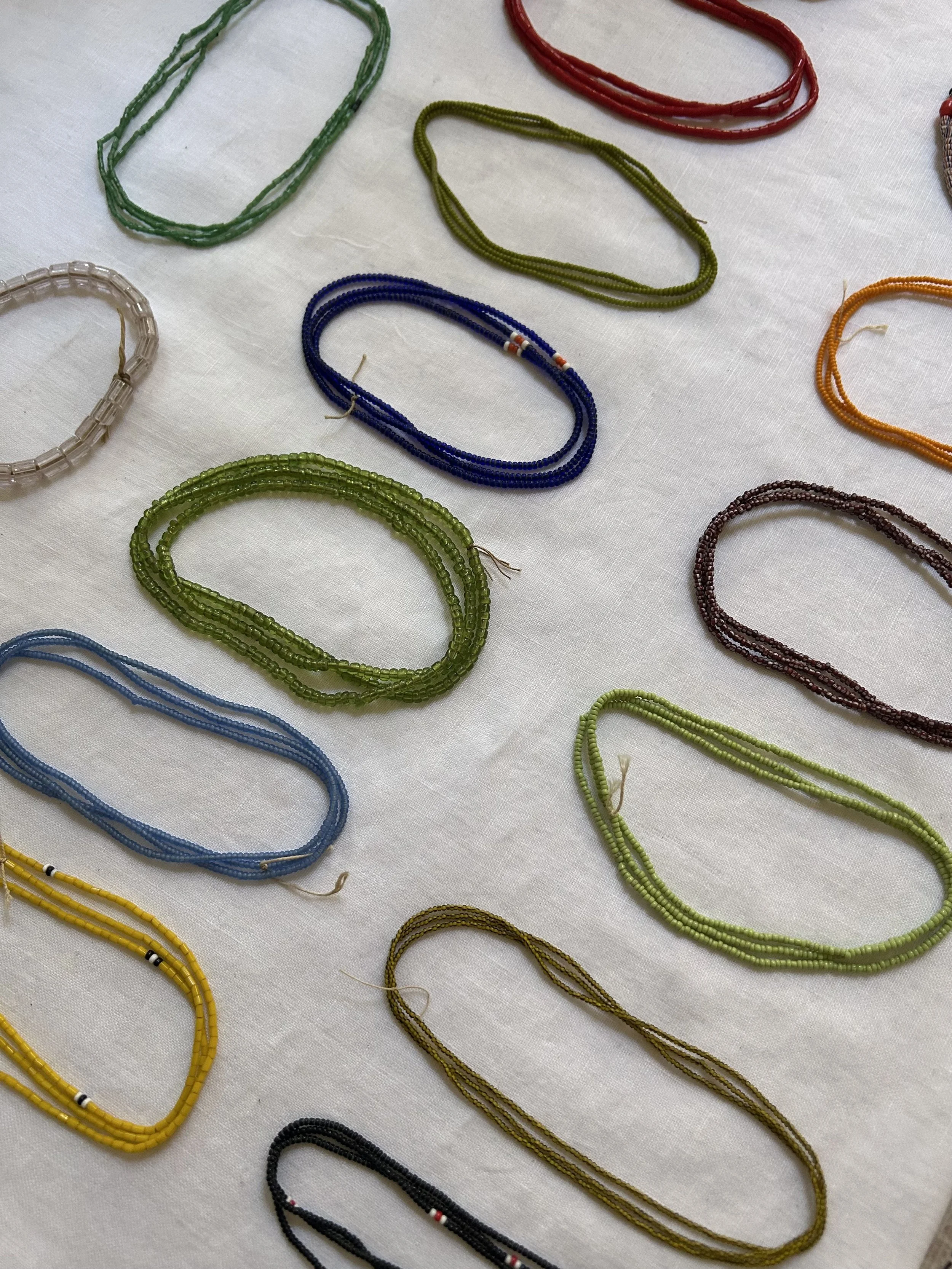Colourful antique beads laid out on a white fabric surface. The necklaces are in various colors including green, blue, red, orange, yellow, black, and white.