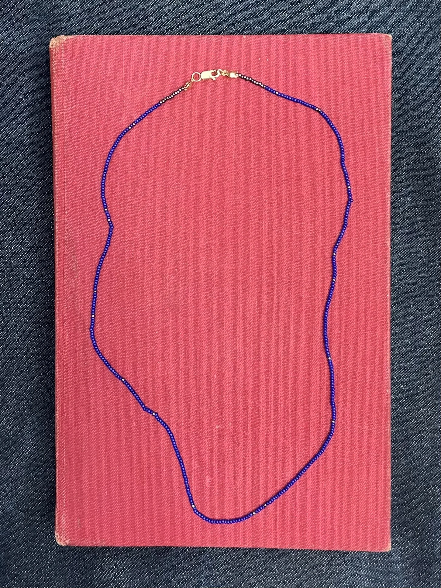 A blue beaded necklace with a gold clasp lies on top of a red hardcover book against a dark fabric background.