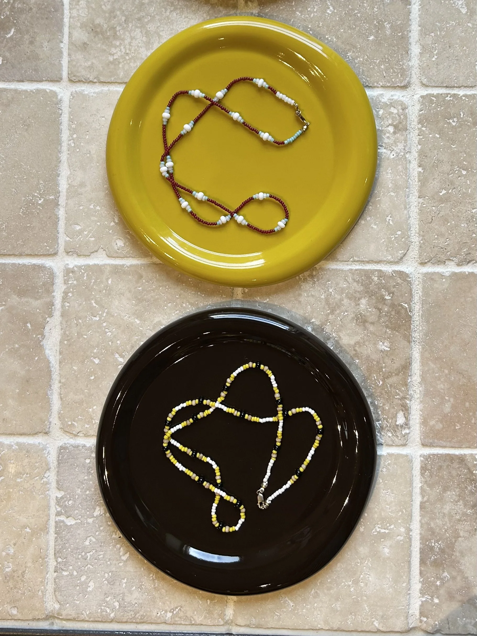Two beaded necklaces, one with white, blue, and red beads on a yellow dish, and the other with black, yellow, and white beads on a black dish made by softedge studio