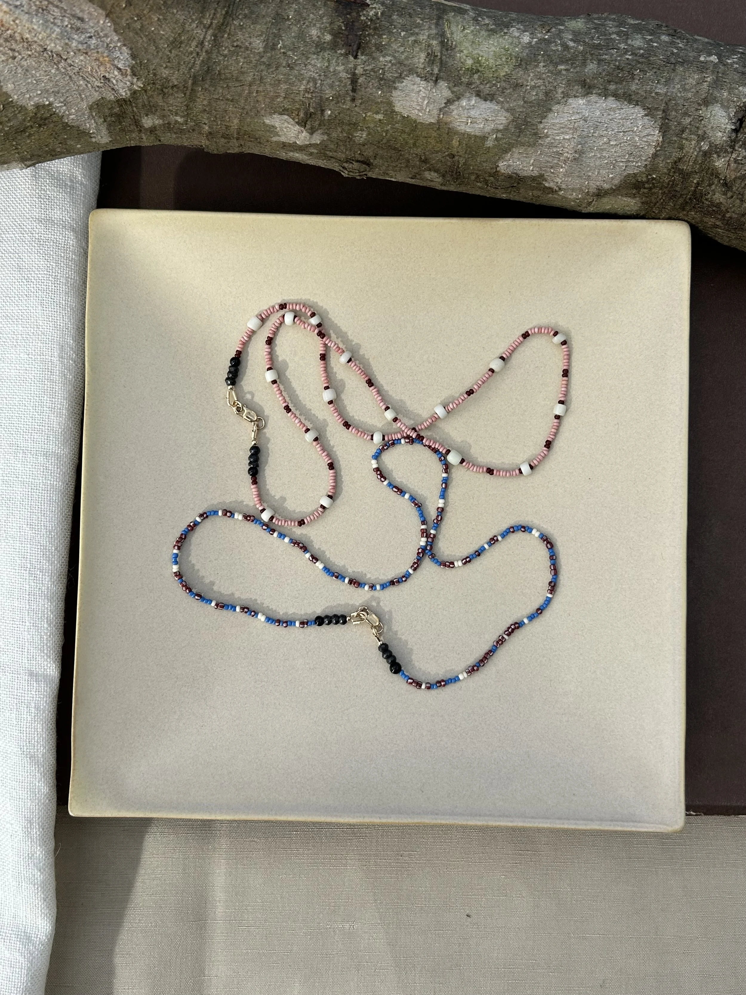Colorful beaded necklaces resting on a beige display tray with a textured surface, next to a piece of raw wood with visible knots and a rough texture.