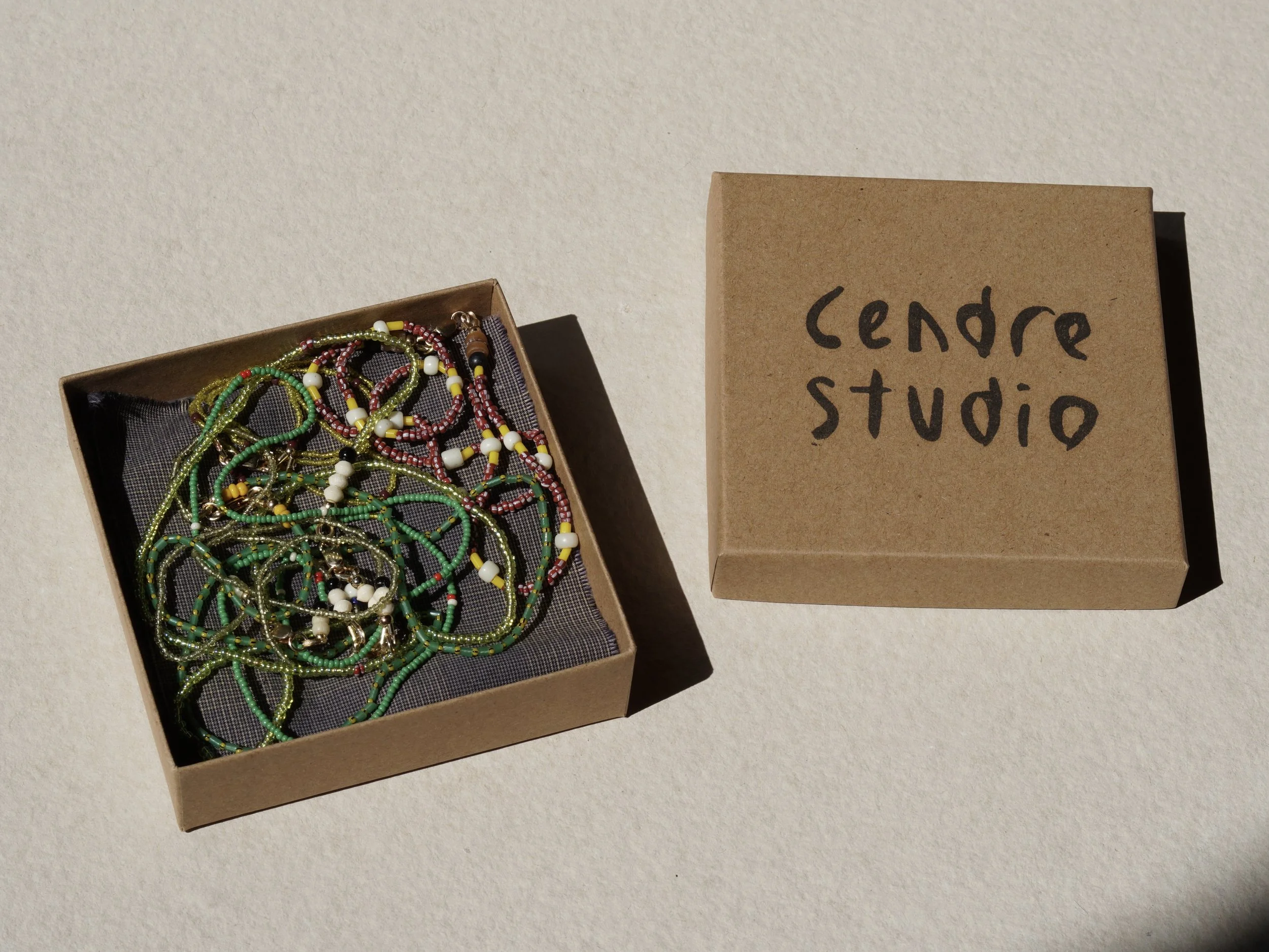 Open cardboard box filled with colorful beaded jewelry, beside a closed cardboard box labeled "Cendre Studio" on a light-colored surface.