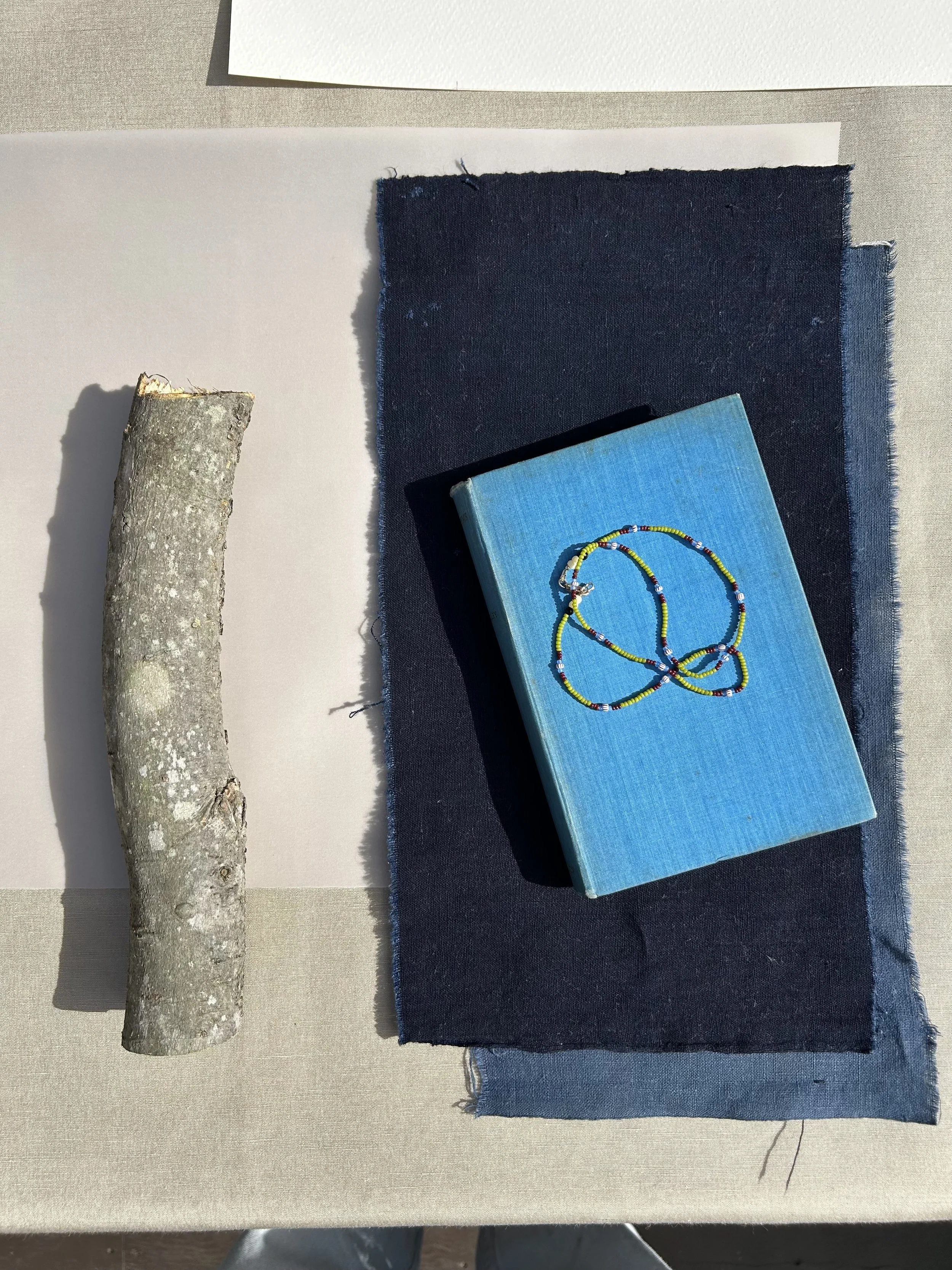 A piece of grey tree branch, a white sheet of paper, a navy blue fabric piece, a light blue book with a beaded necklace on top, and a light-colored surface.