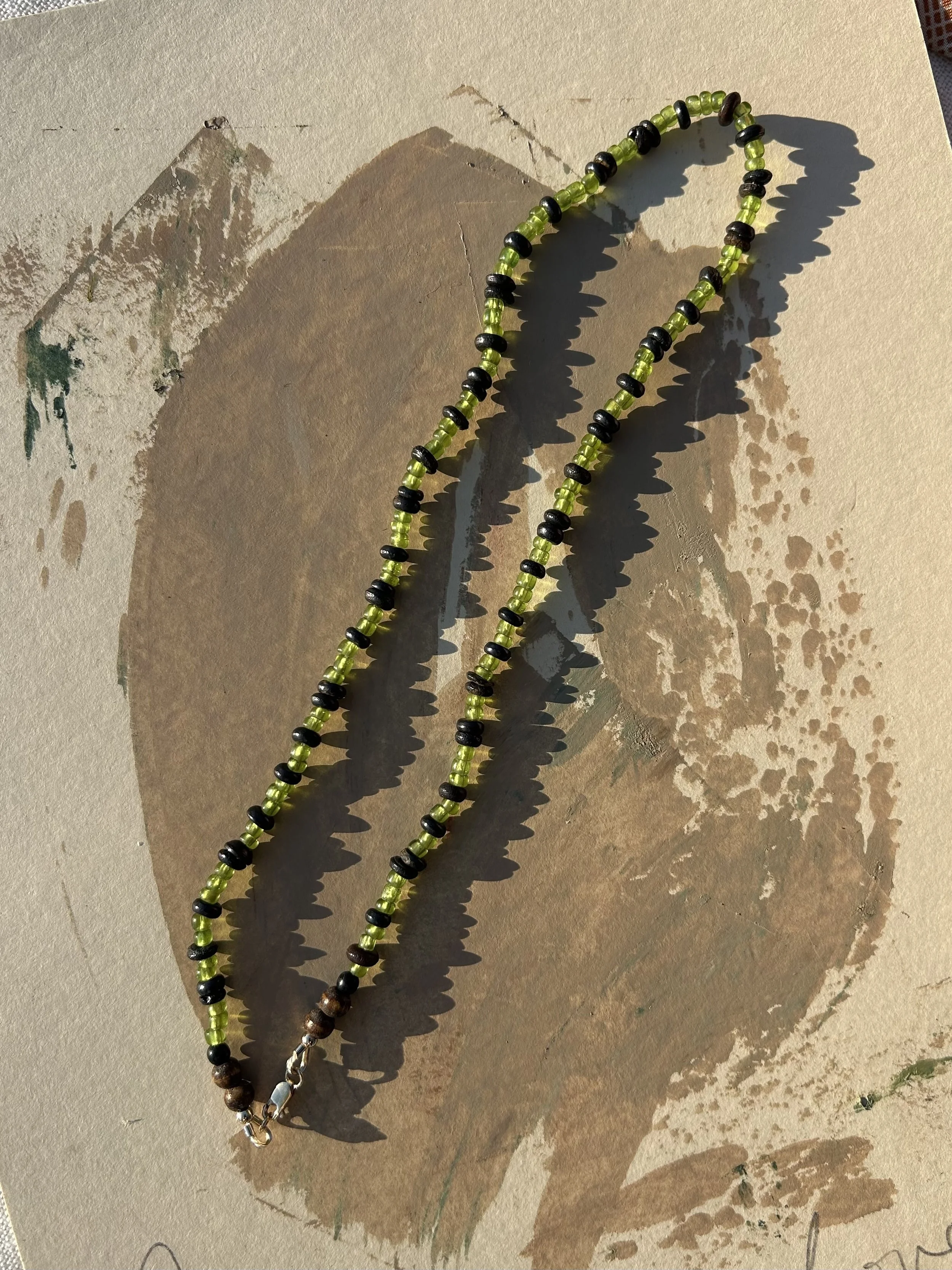 Two beaded necklaces with green and black beads, laid over a painting in shades of brown and beige by Max Ososki