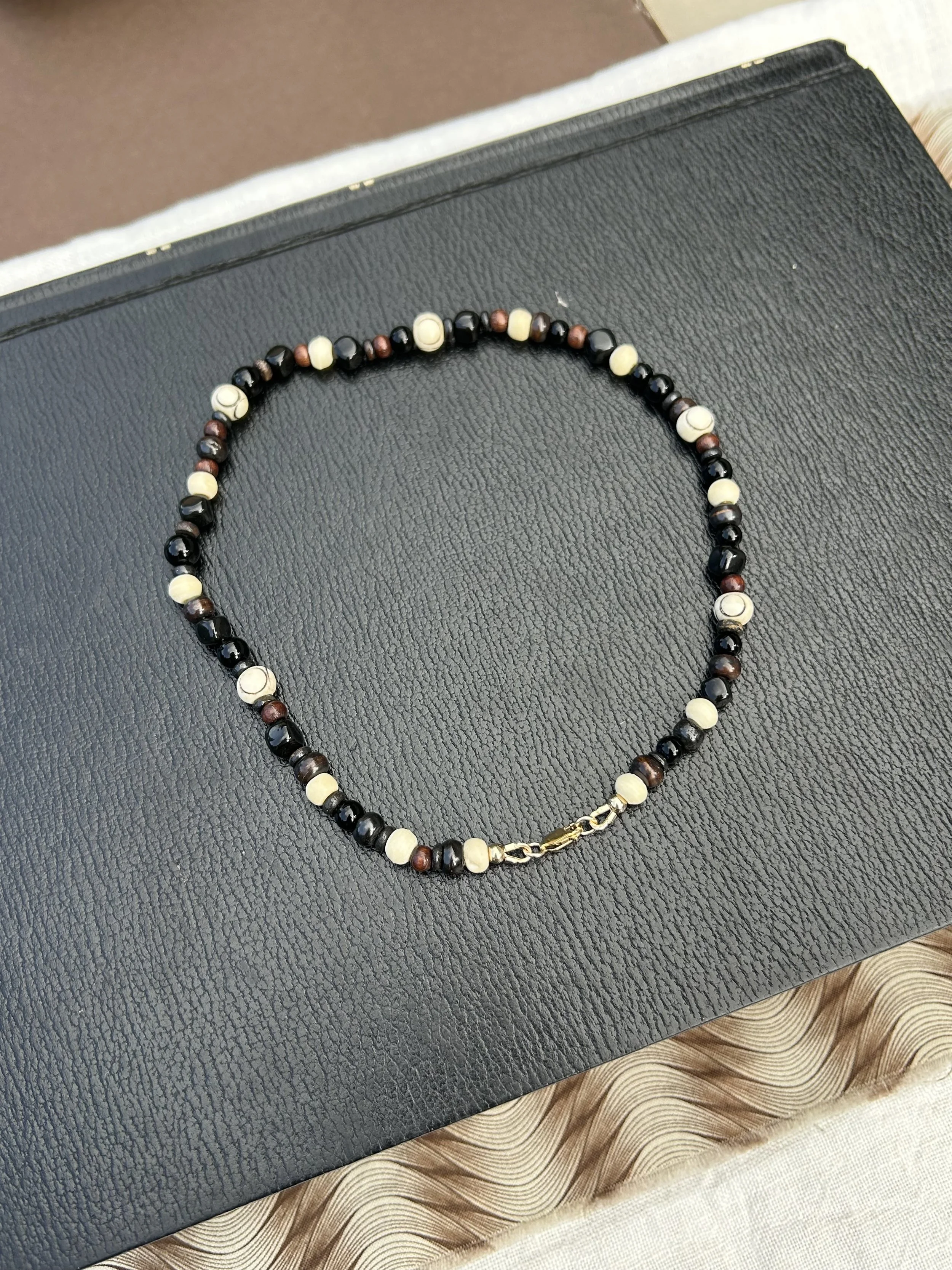 A beaded necklace with alternating black, brown, and cream-colored beads, featuring a gold-colored clasp, resting on a black textured surface with a beige and brown decorative cloth underneath.
