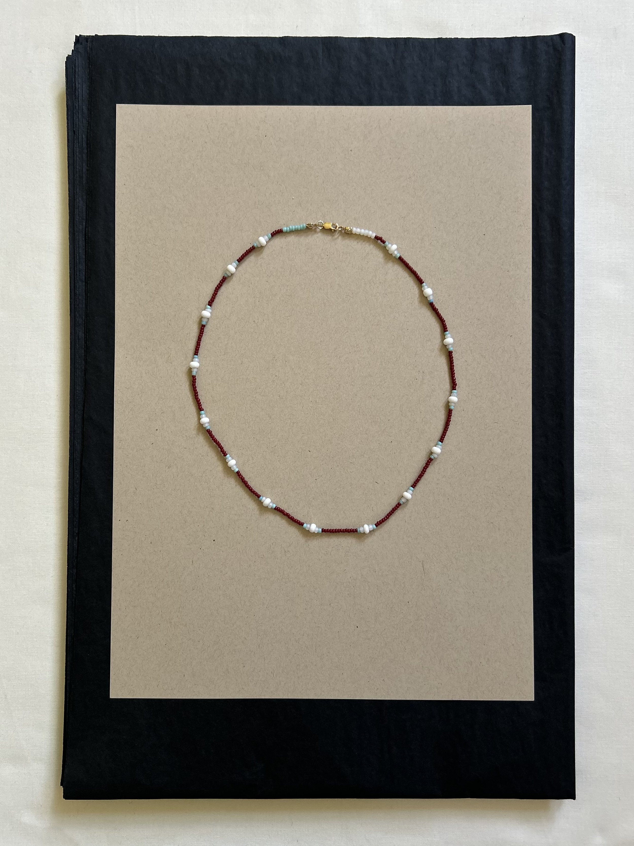 Beaded necklace with red, white, and light blue beads arranged in a pattern, lying on a beige paper sheet on top of a black hardcover book.