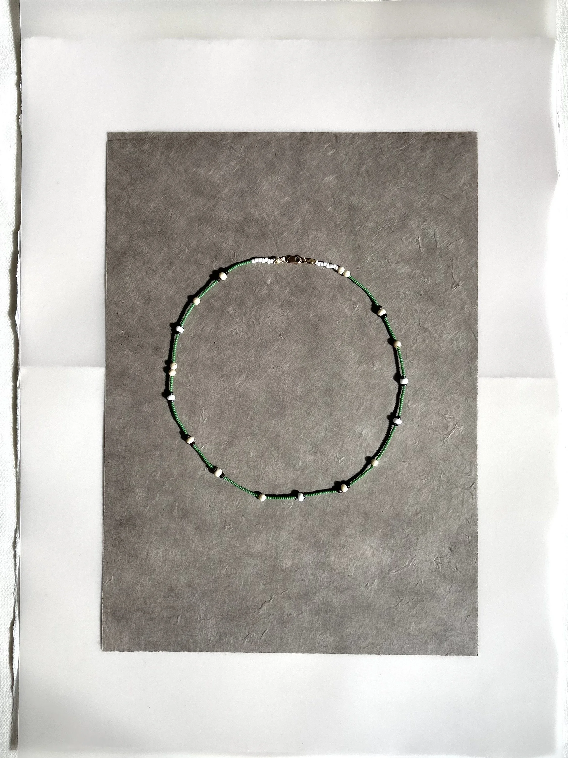 A beaded necklace with green, white, and black beads, displayed on a gray background with white paper border.